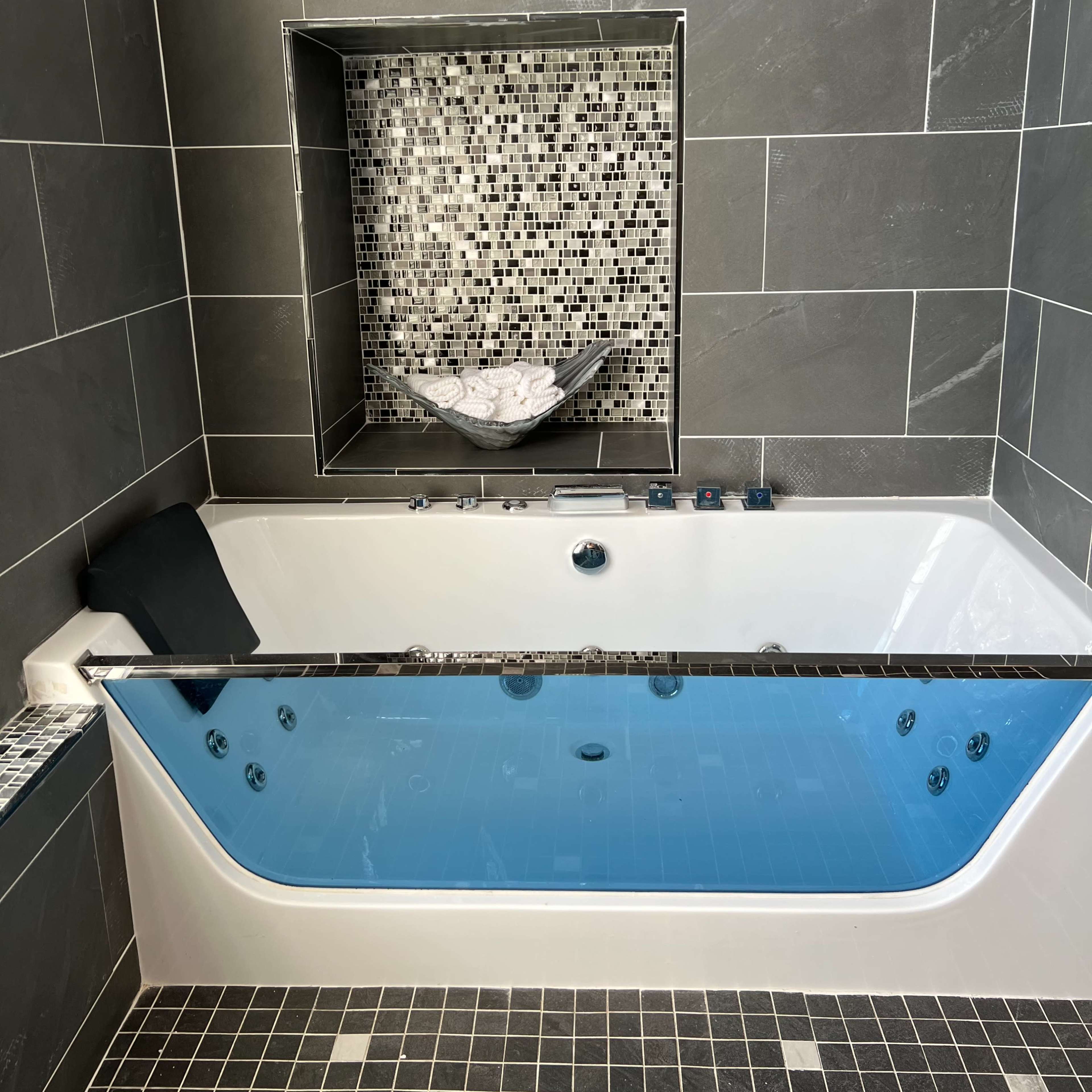 A modern bathtub with an inset shelf, surrounded by dark tiled walls and a mosaic accent, sits against the backdrop of a window.
