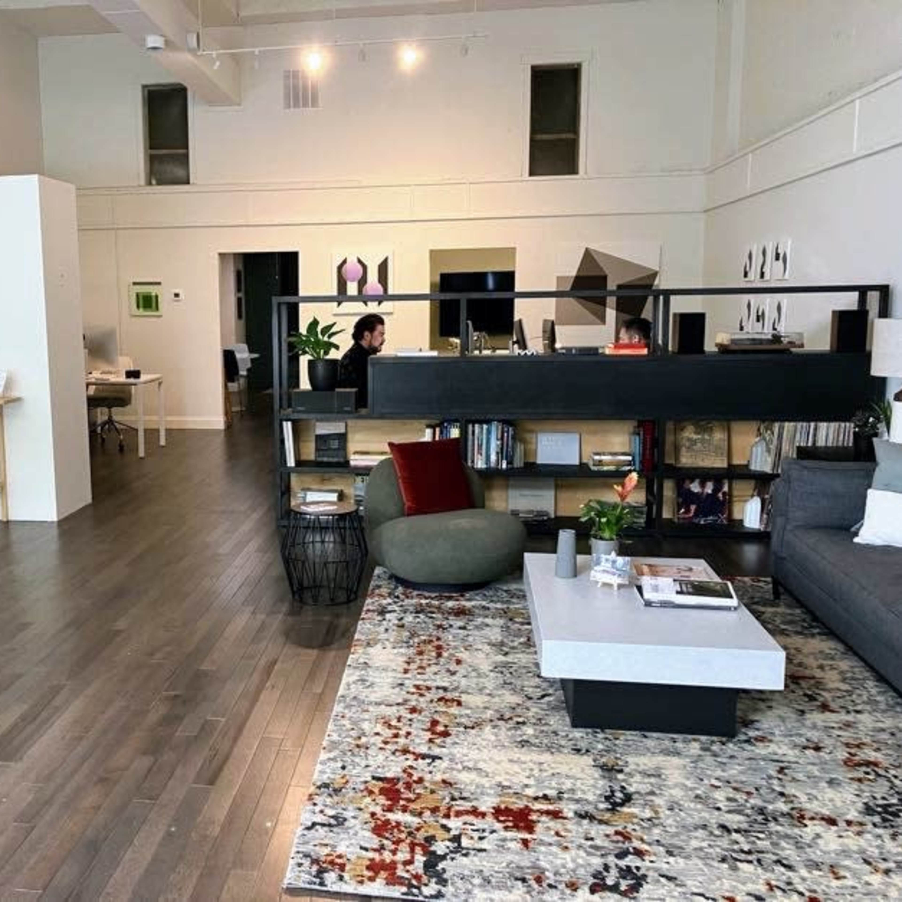 A spacious modern living area featuring a gray sofa, a coffee table on a patterned rug, a bookshelf, and distinct workspaces in the background.