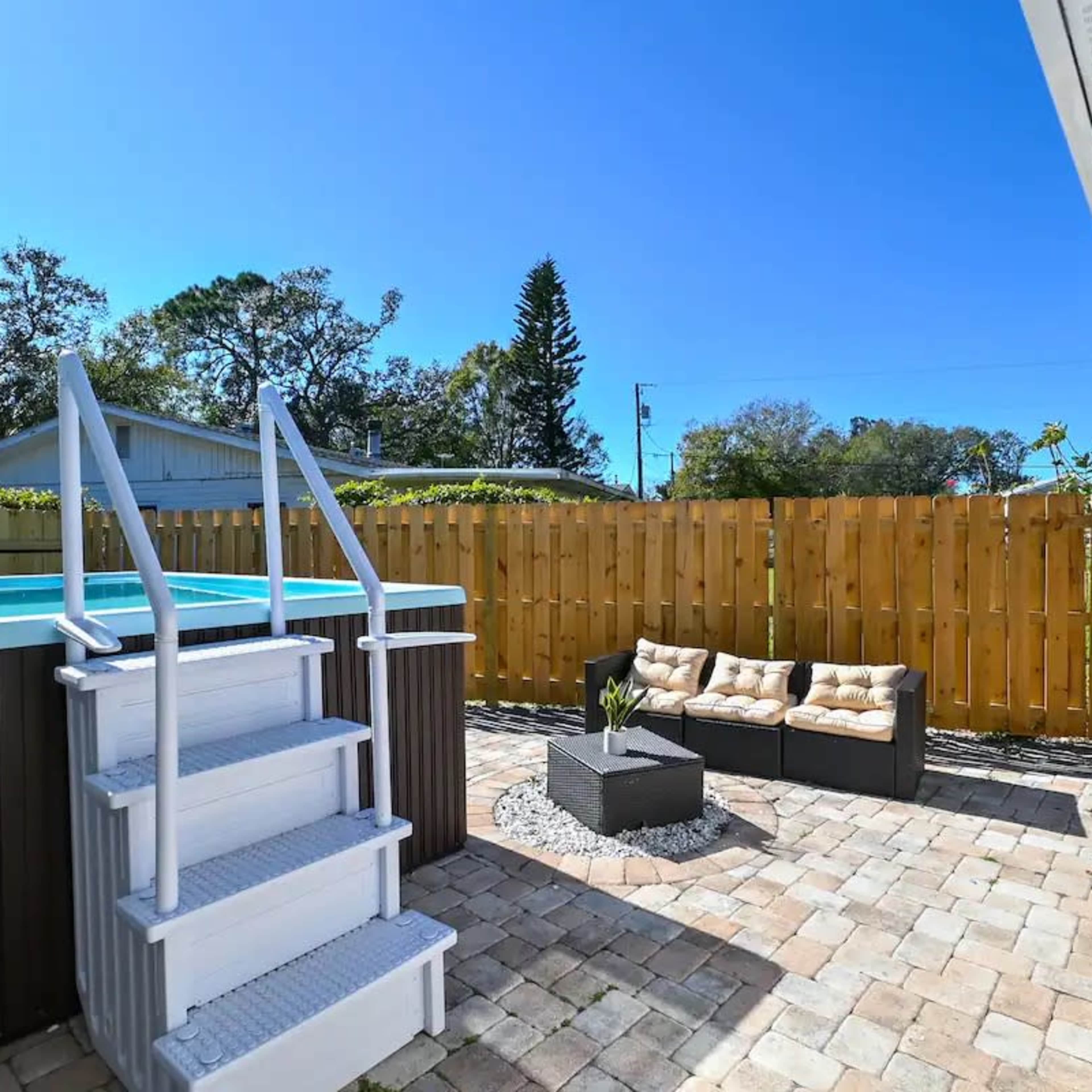 A small outdoor patio with a wooden fence, a raised pool, and a seating area made of black wicker furniture with cushions.