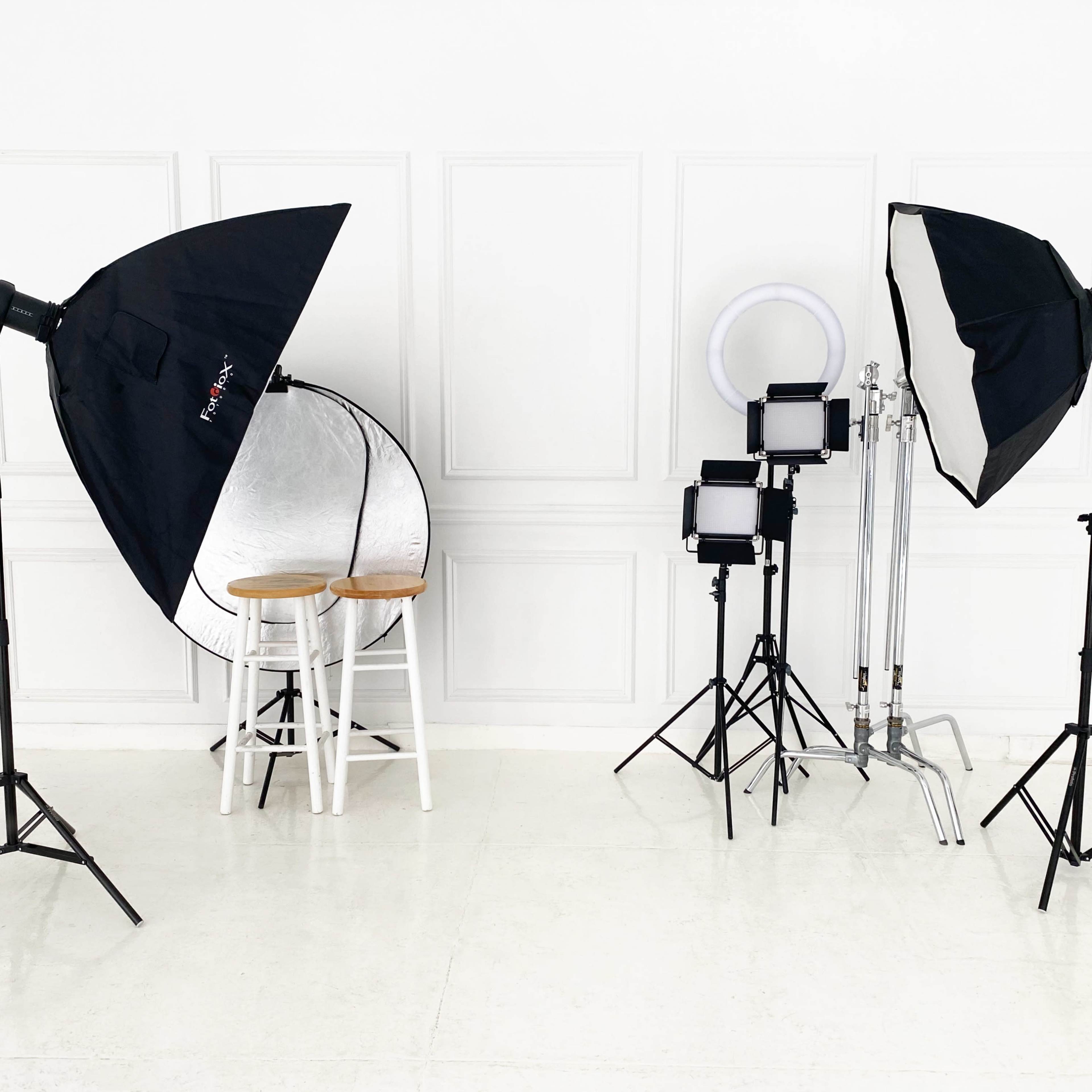 A photography studio setup with lighting equipment, a round reflector, and two stools positioned on a white backdrop.