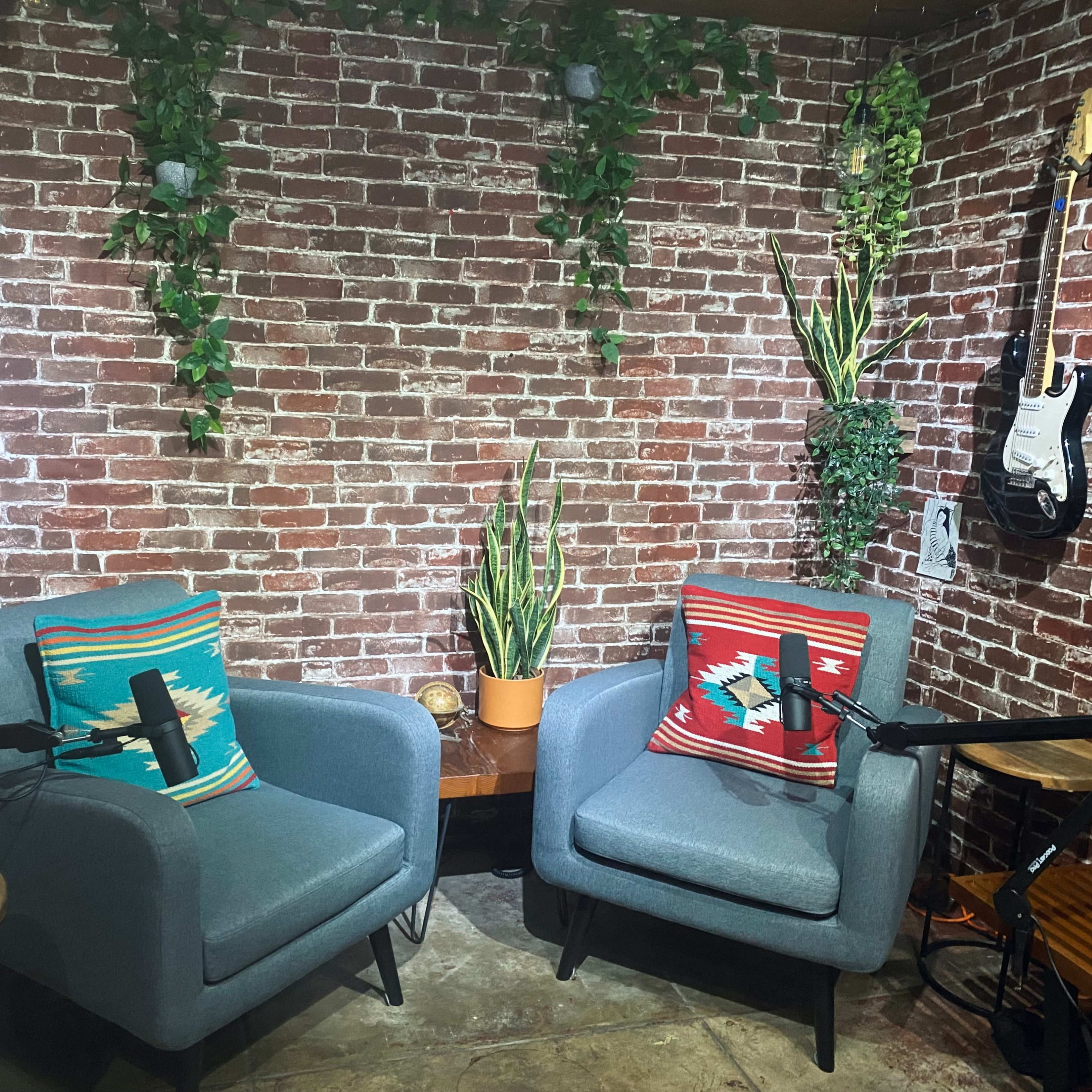 Two blue armchairs with colorful pillows positioned in front of a brick wall, accompanied by plants and a guitar on display.