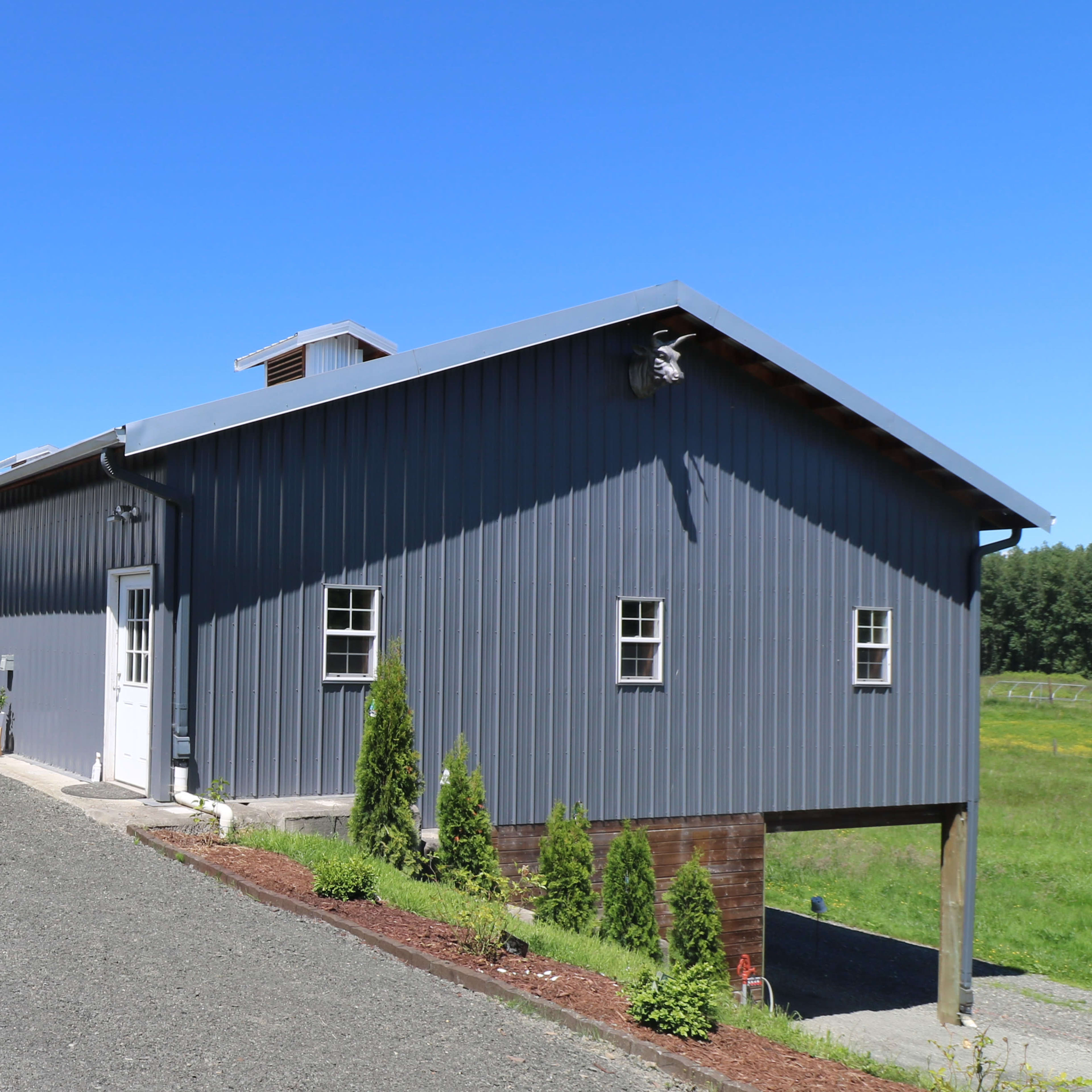 A metal-clad building with a gable roof is situated beside a gravel driveway, surrounded by grassy fields.