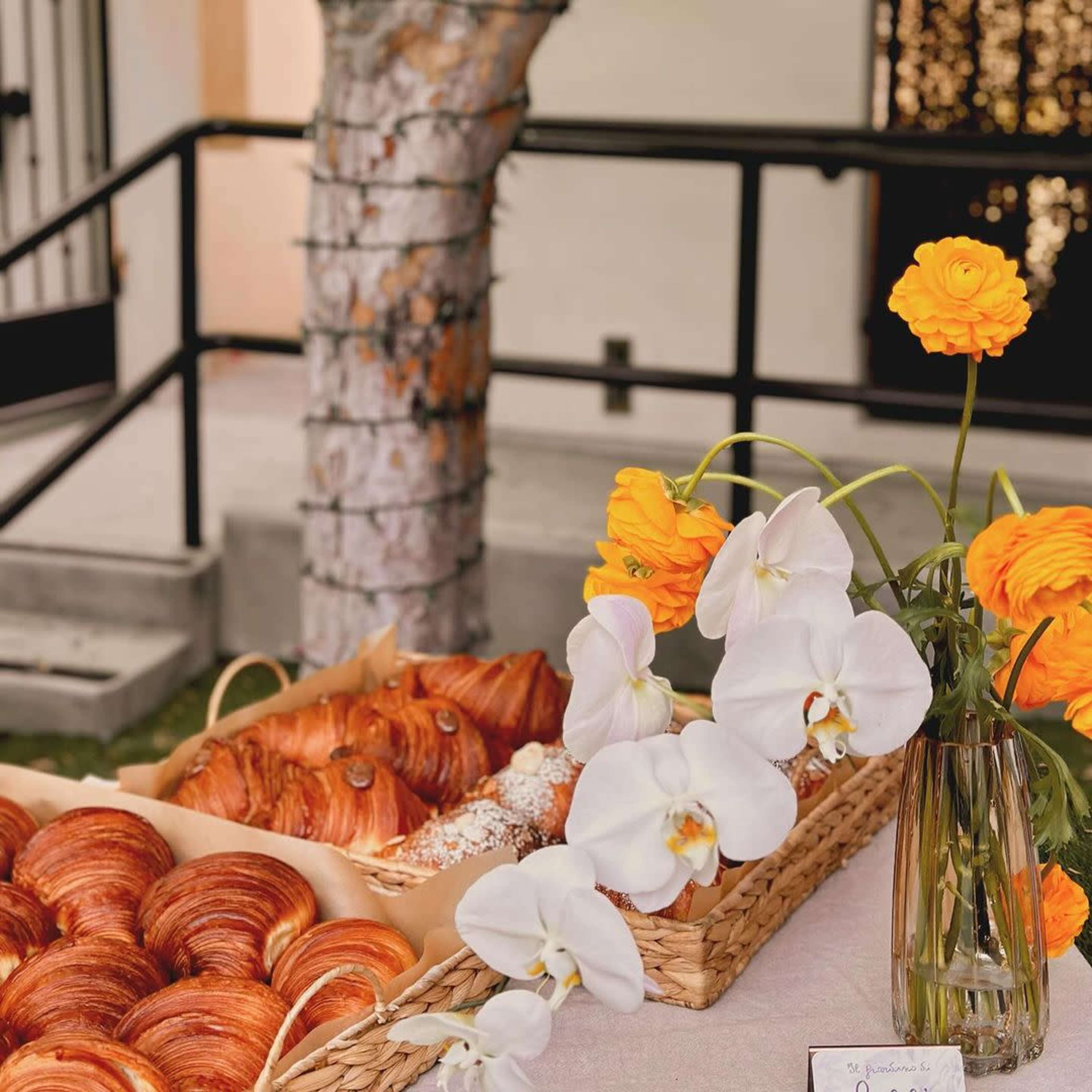 A table displays a variety of croissants and pastries alongside a vase of white orchids and bright orange ranunculus flowers.