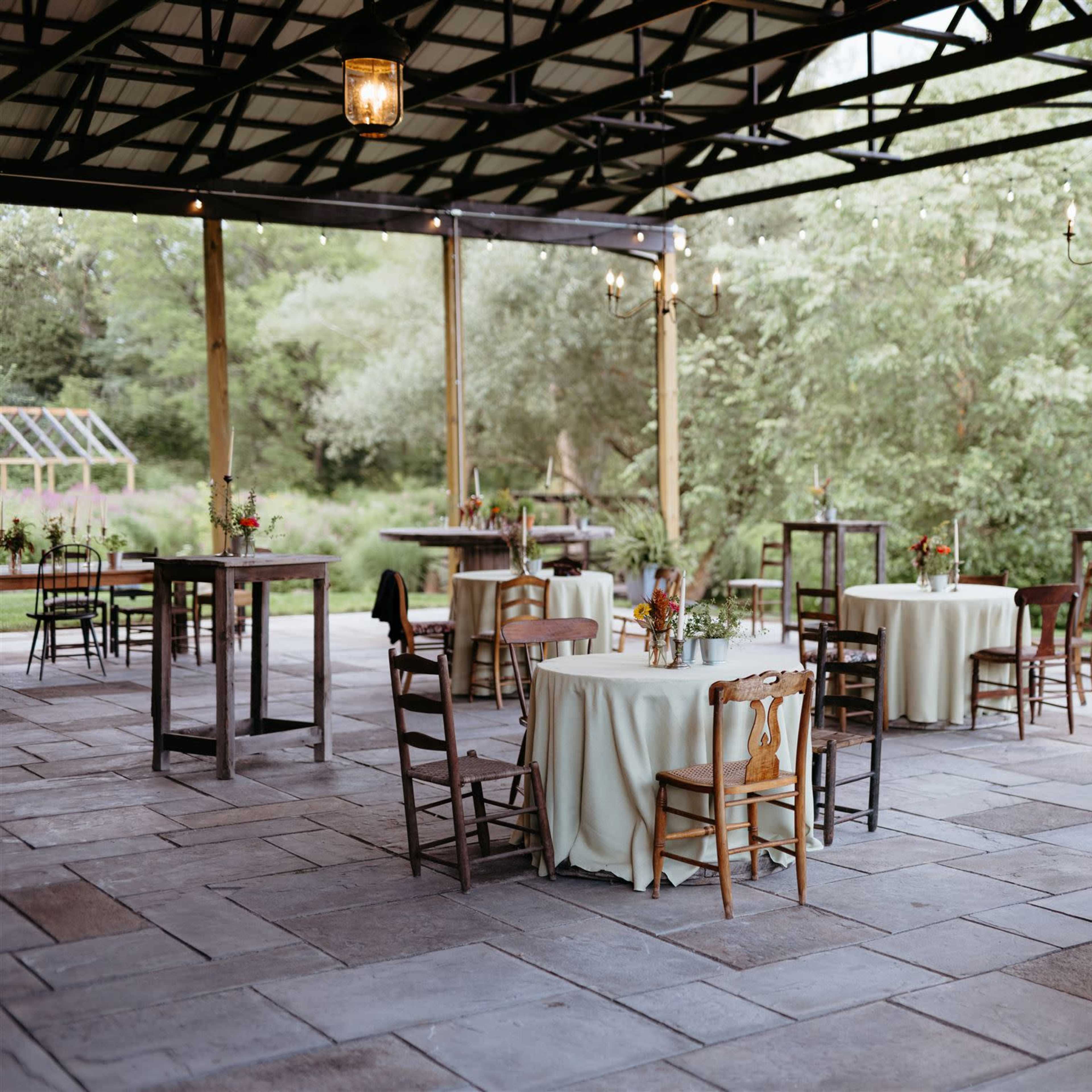 A spacious outdoor pavilion features several tables with light-colored tablecloths arranged on a stone floor, surrounded by greenery.