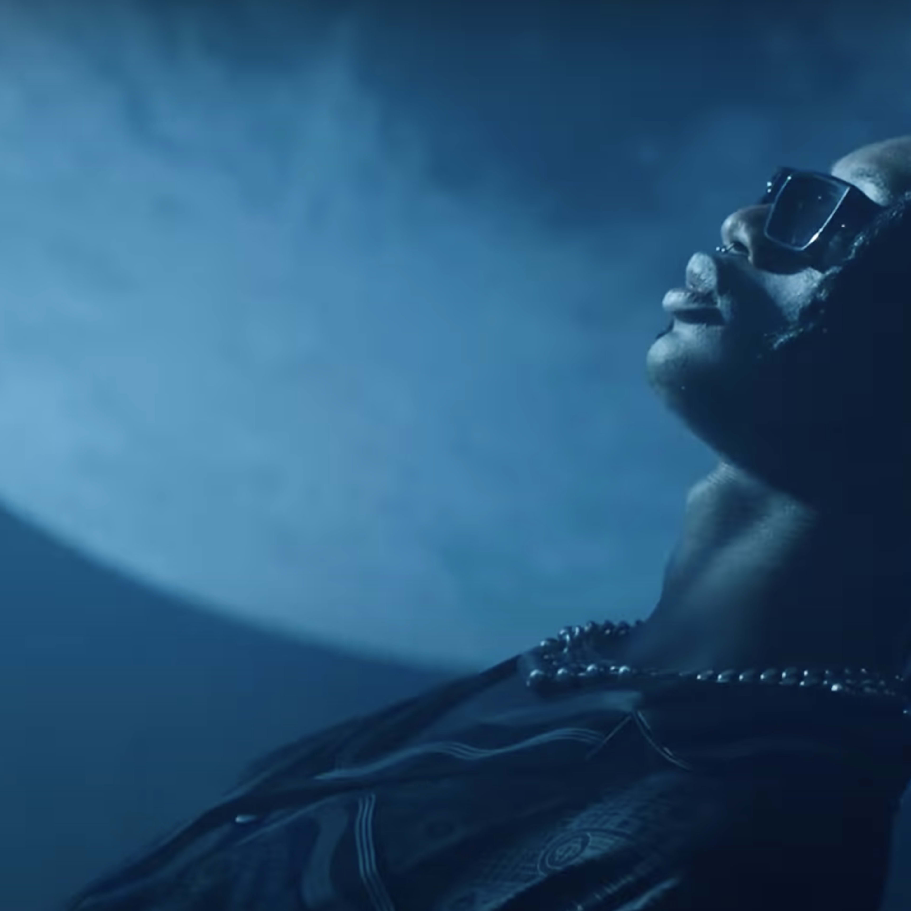 A person wearing sunglasses and a necklace poses against a large, illuminated moon in the background.