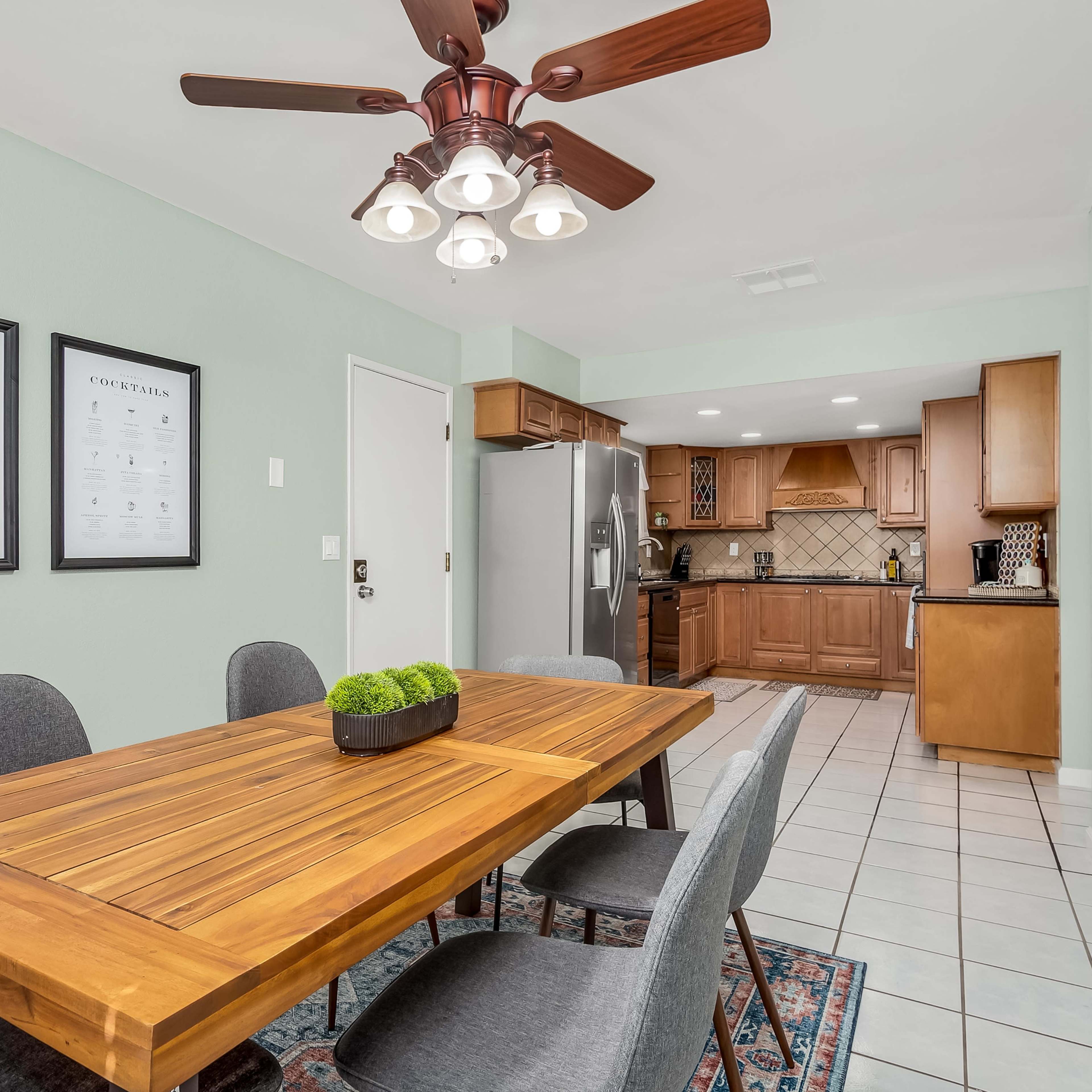 A modern dining area features a wooden table surrounded by gray chairs, with a ceiling fan overhead and an adjoining kitchen visible in the background.