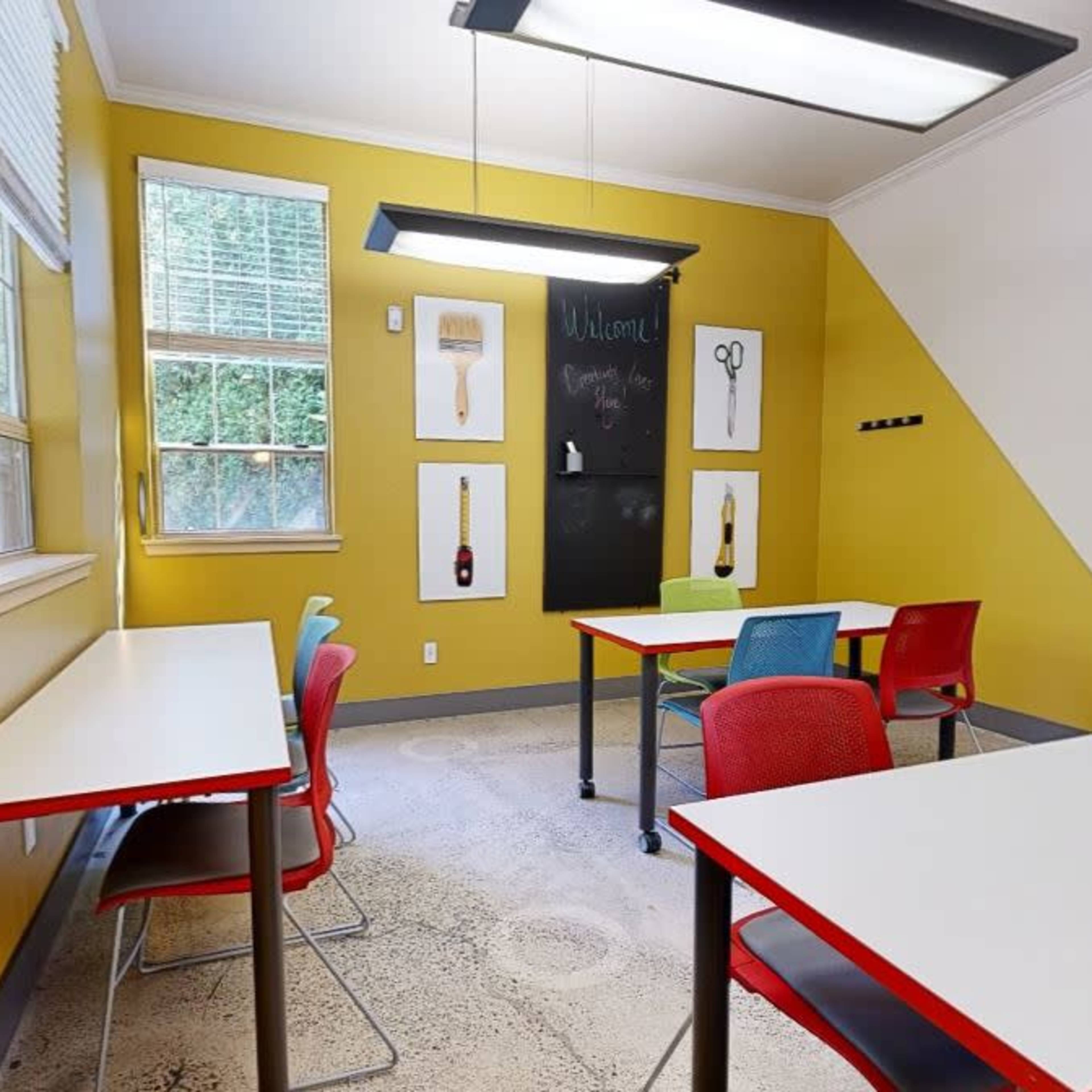 A classroom features bright yellow walls, two tables with red and gray chairs, and artwork hanging on the walls.