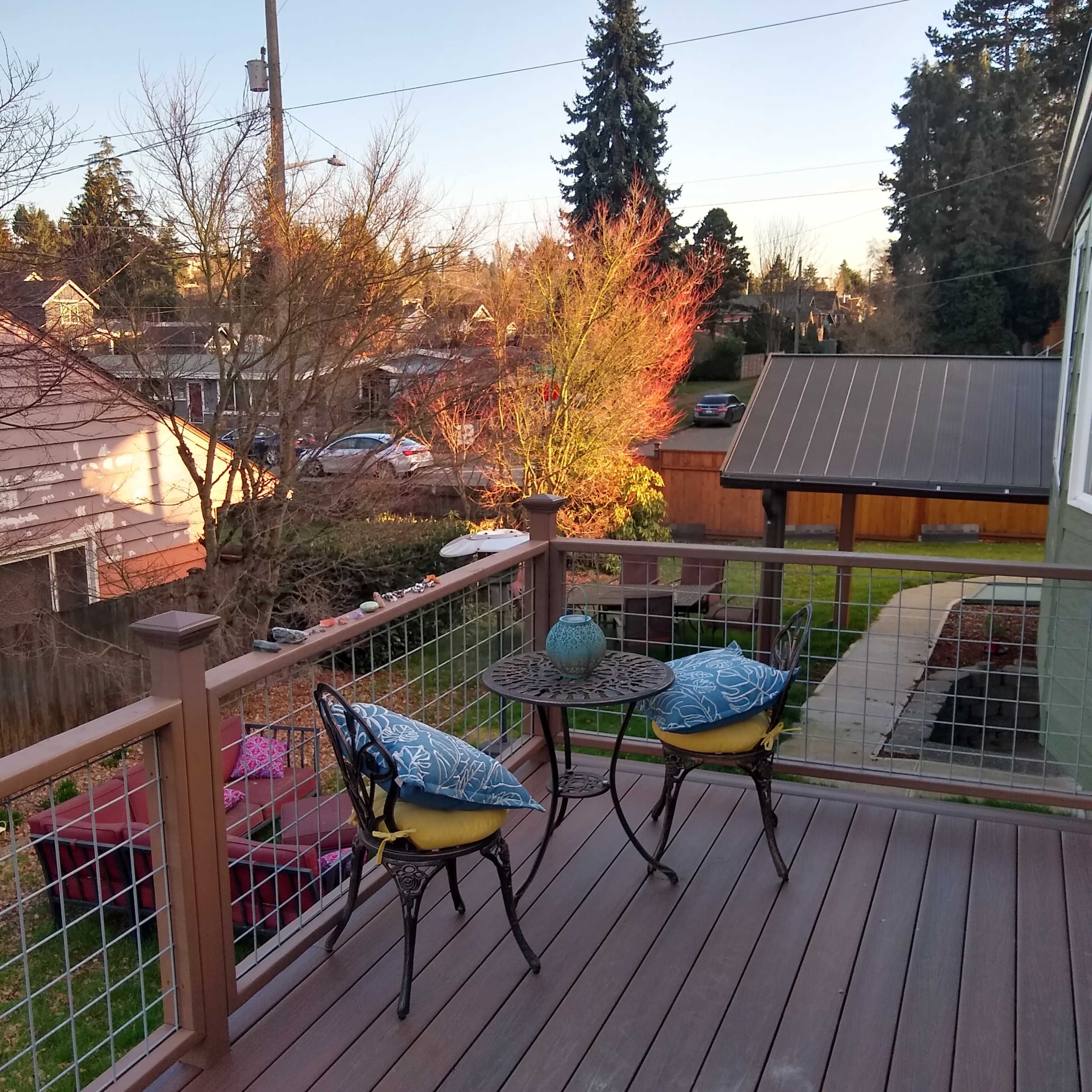 A wooden deck with two cushioned chairs and a small table, overlooking a landscaped backyard and trees in the distance.