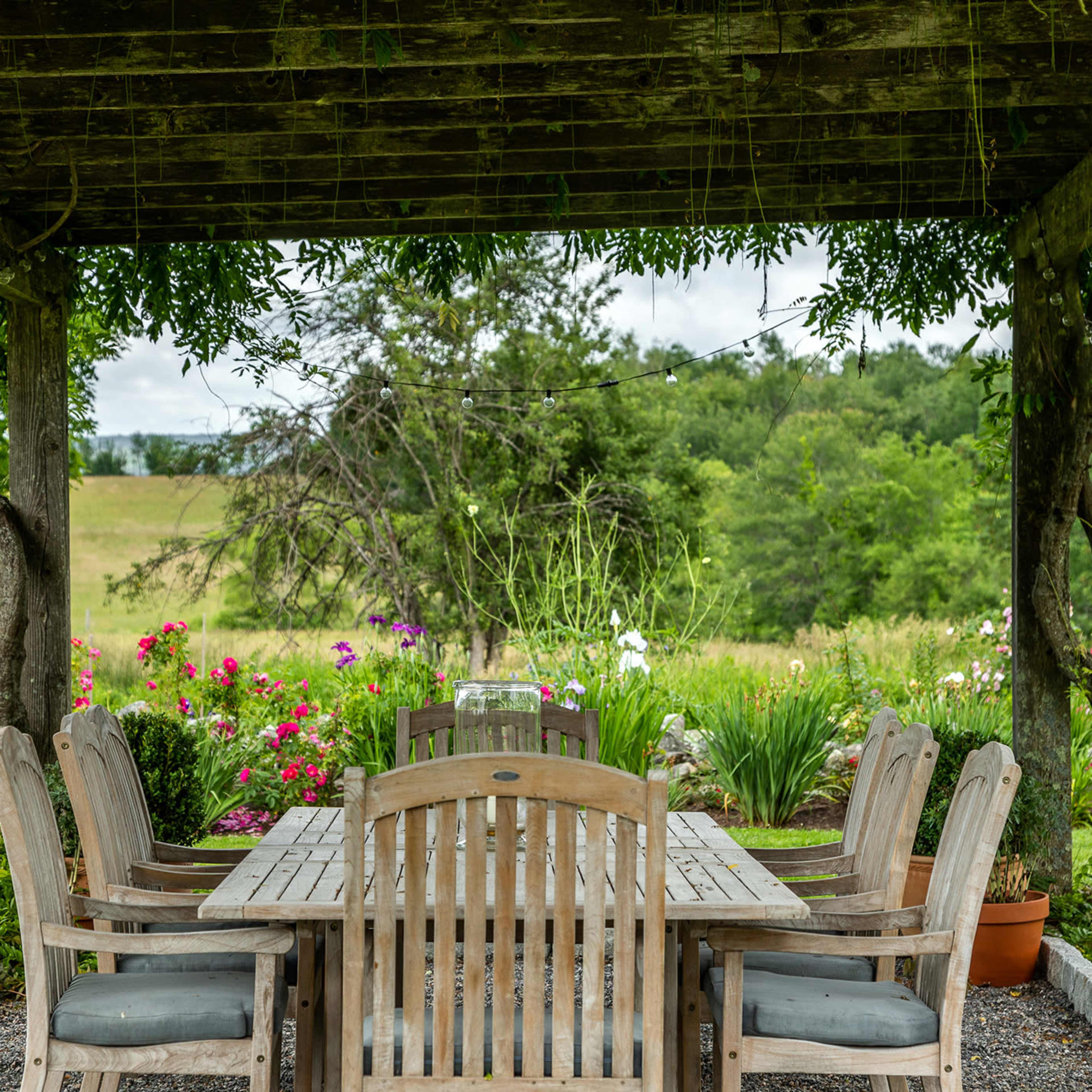 A wooden dining table surrounded by chairs is set under a rustic pergola, with a backdrop of lush greenery and blooming flowers.