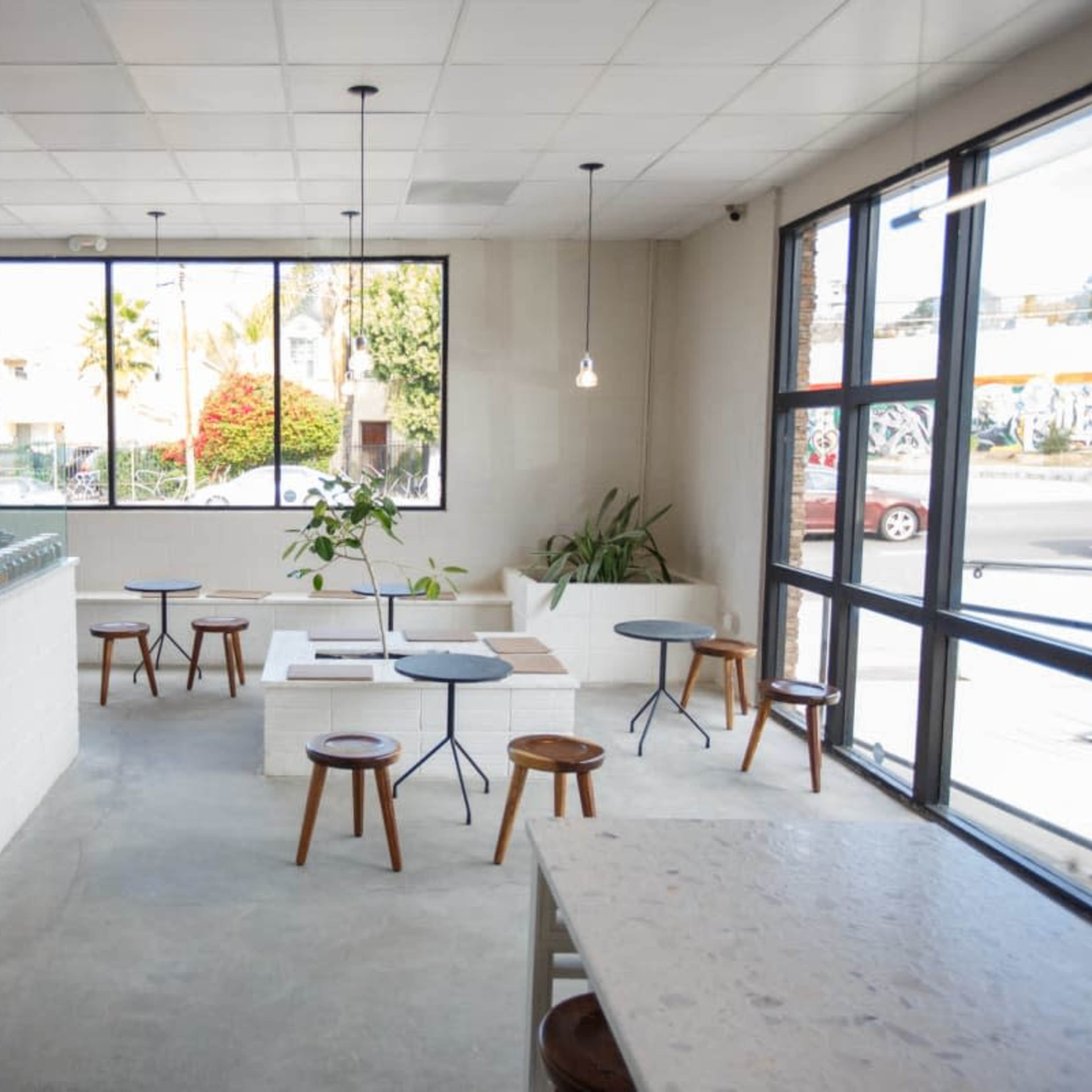 A modern café interior with minimal furnishings, featuring a counter on the left, small round tables, and ample natural light from large windows.
