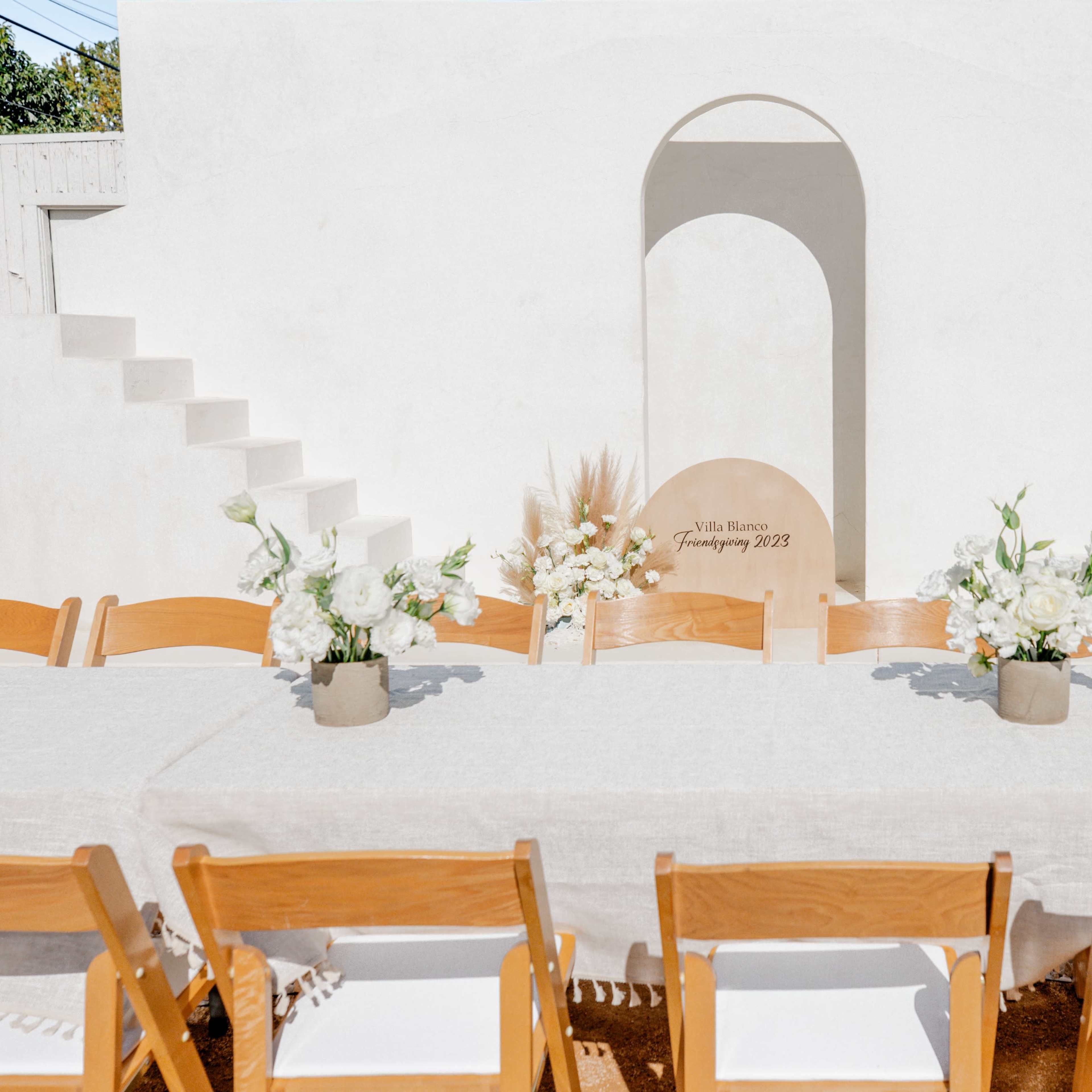 A long table adorned with floral centerpieces is set up in front of a minimalist white wall featuring an arch and staircase.