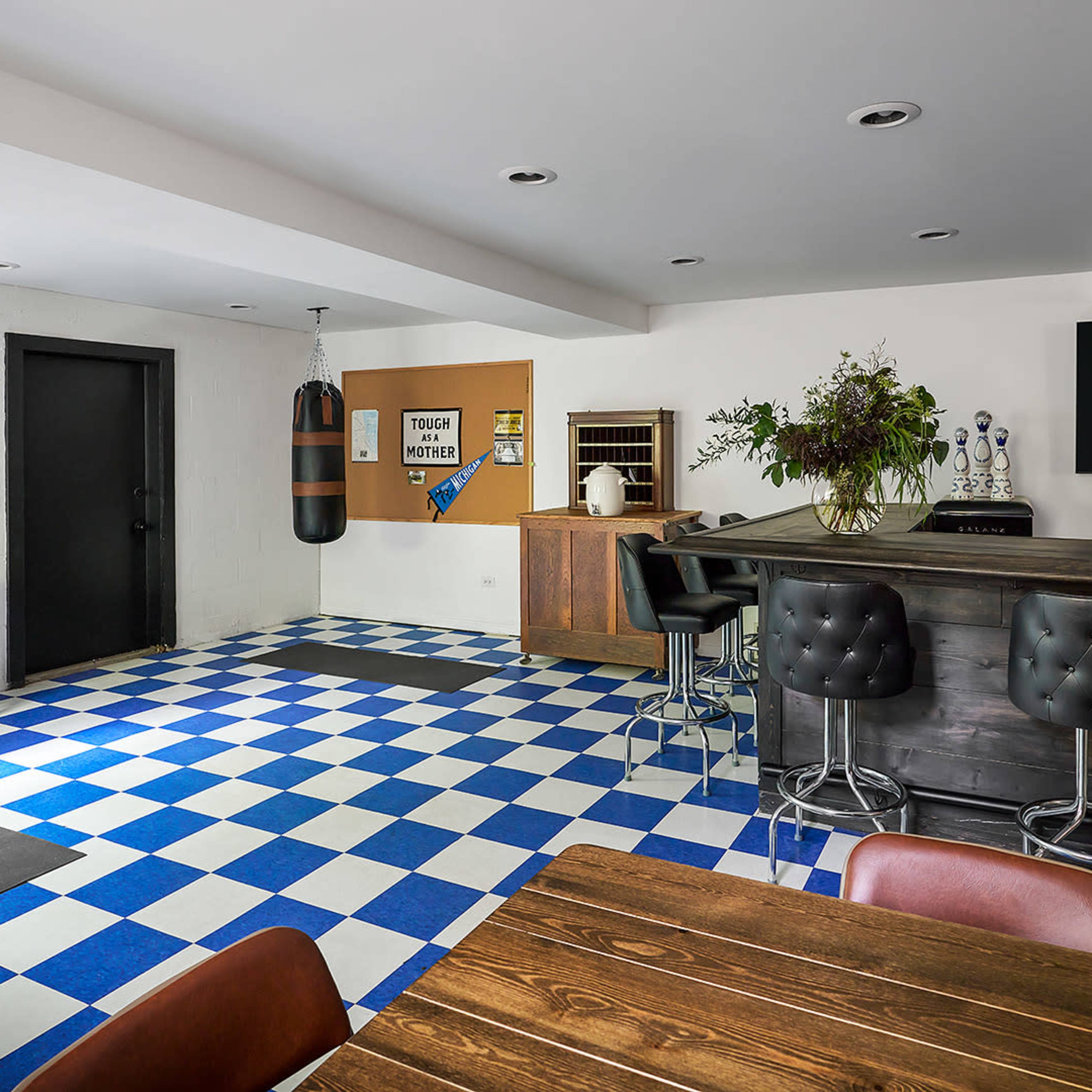 A basement room featuring a boxing bag, a bar area, a dining table, and a checkered blue and white tiled floor.