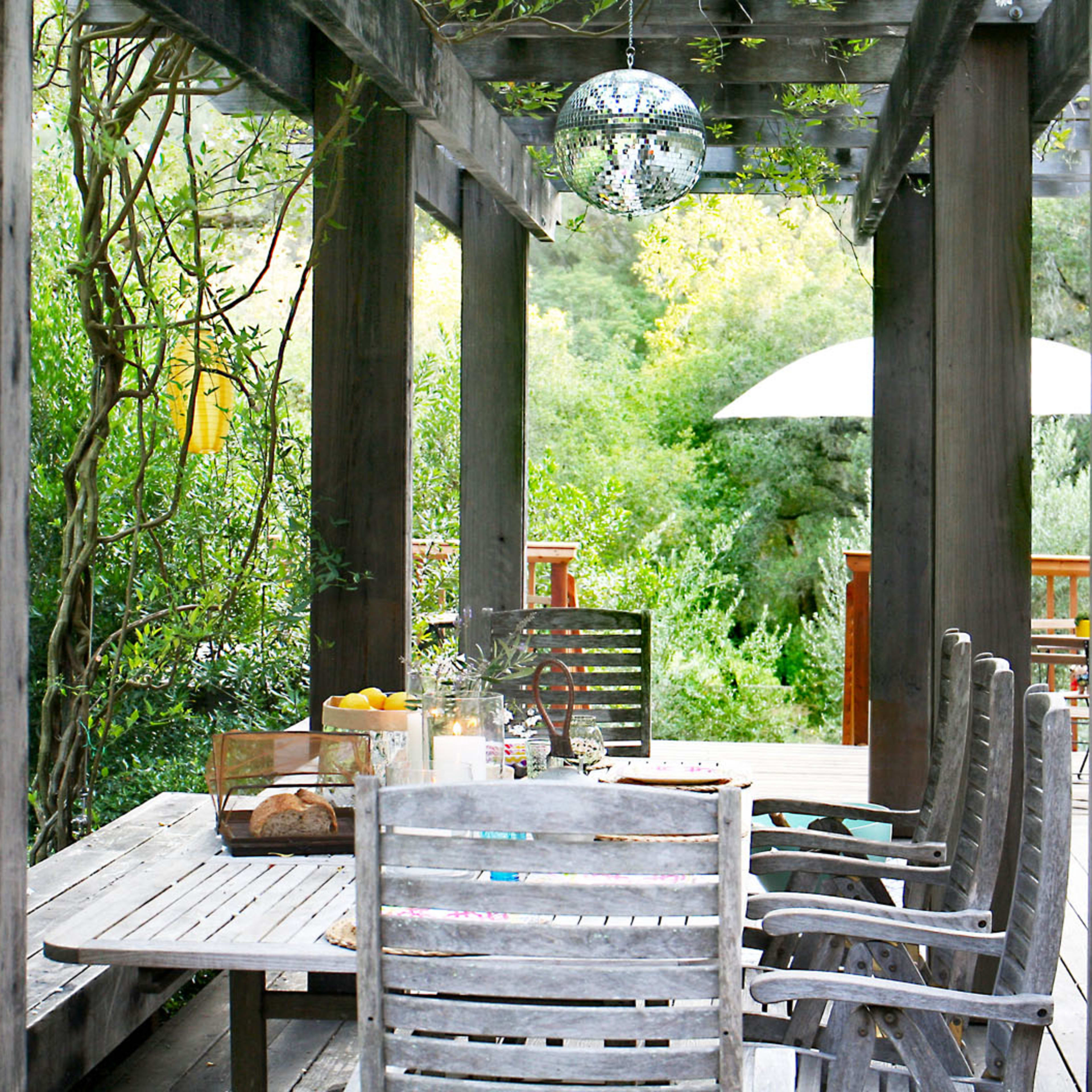 A wooden table with several chairs is set under a pergola surrounded by greenery and illuminated by a hanging disco ball.