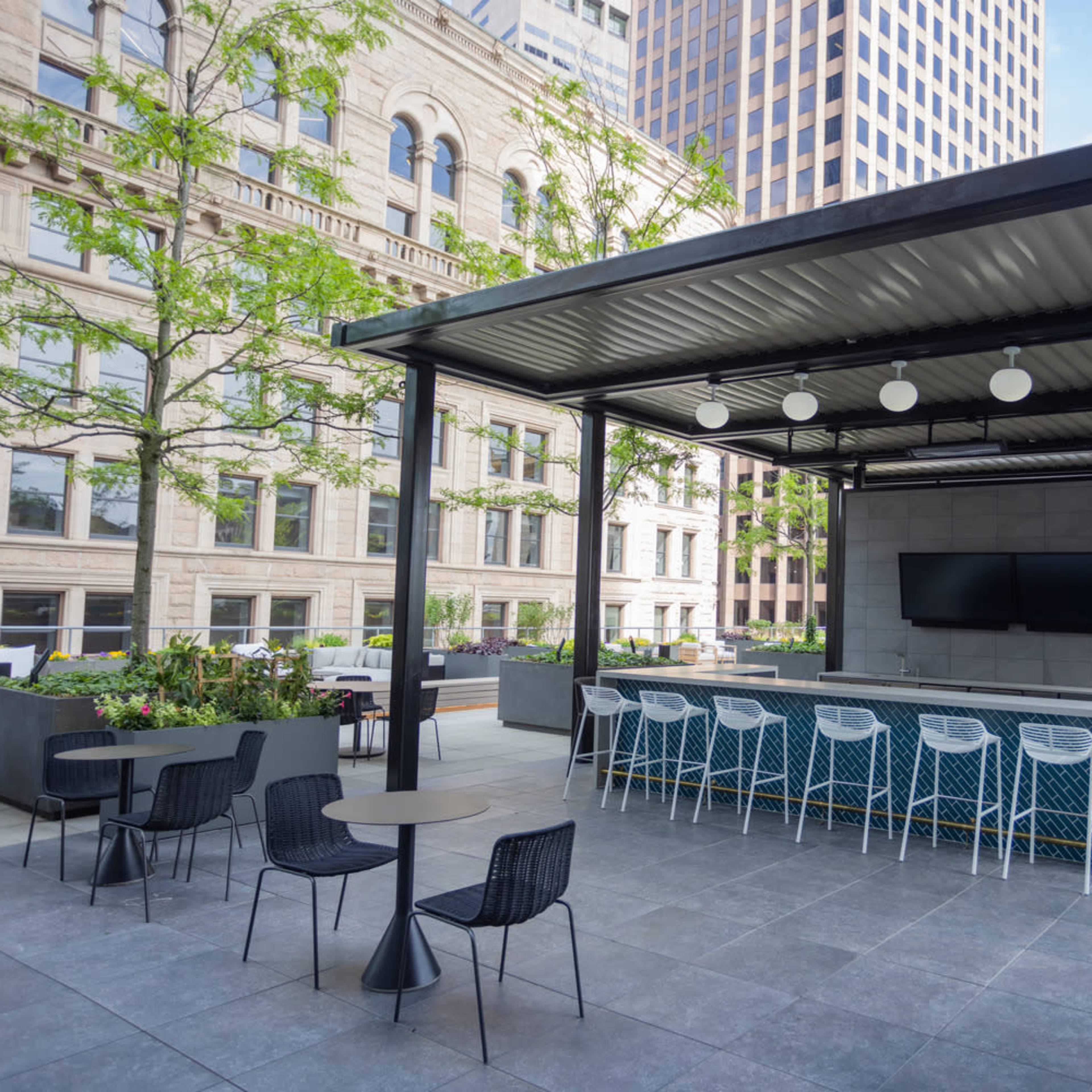 An outdoor patio area with a covered bar, seating, and two television screens, surrounded by modern landscaping and buildings.