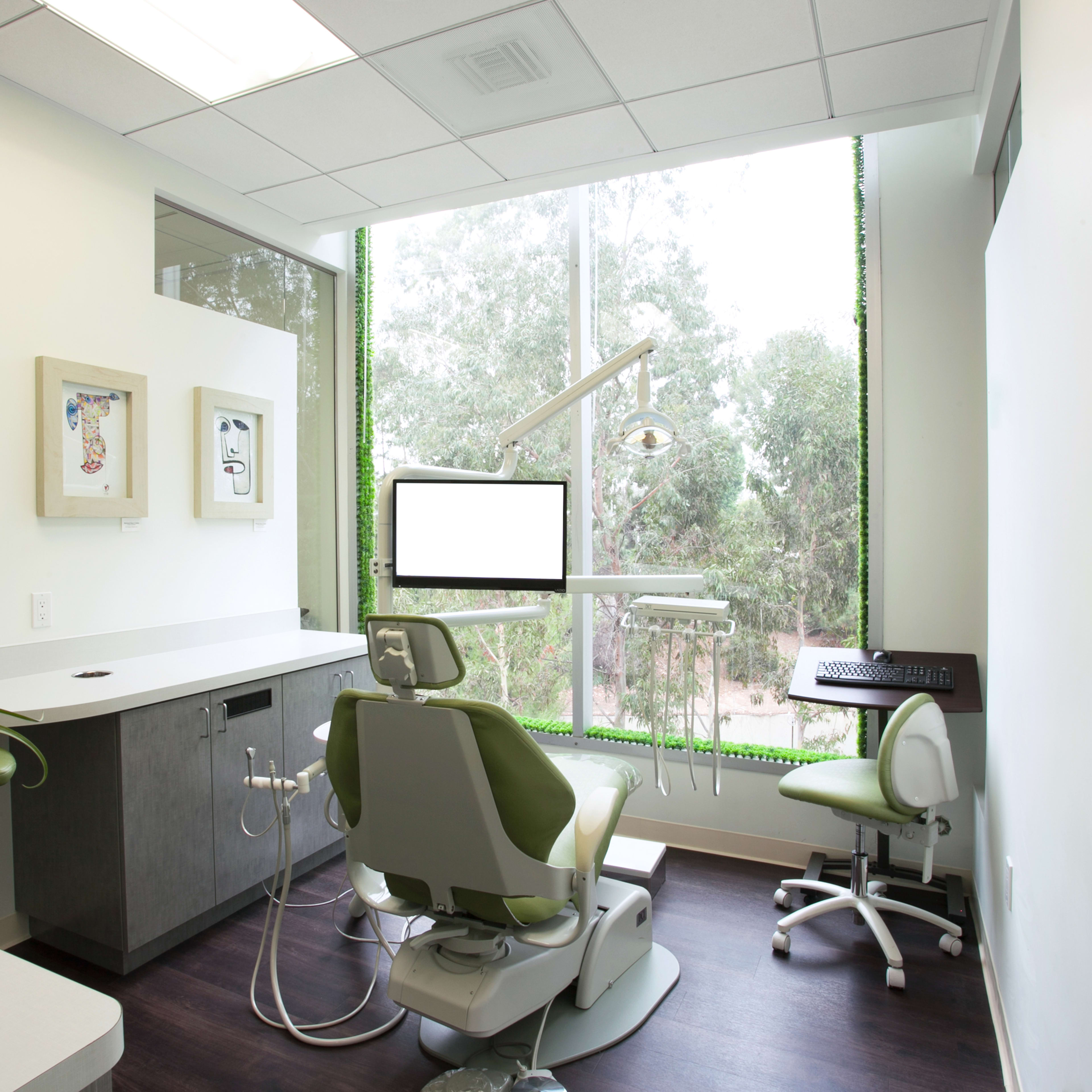 A dental office features a treatment chair and equipment positioned near a large window with greenery visible outside.