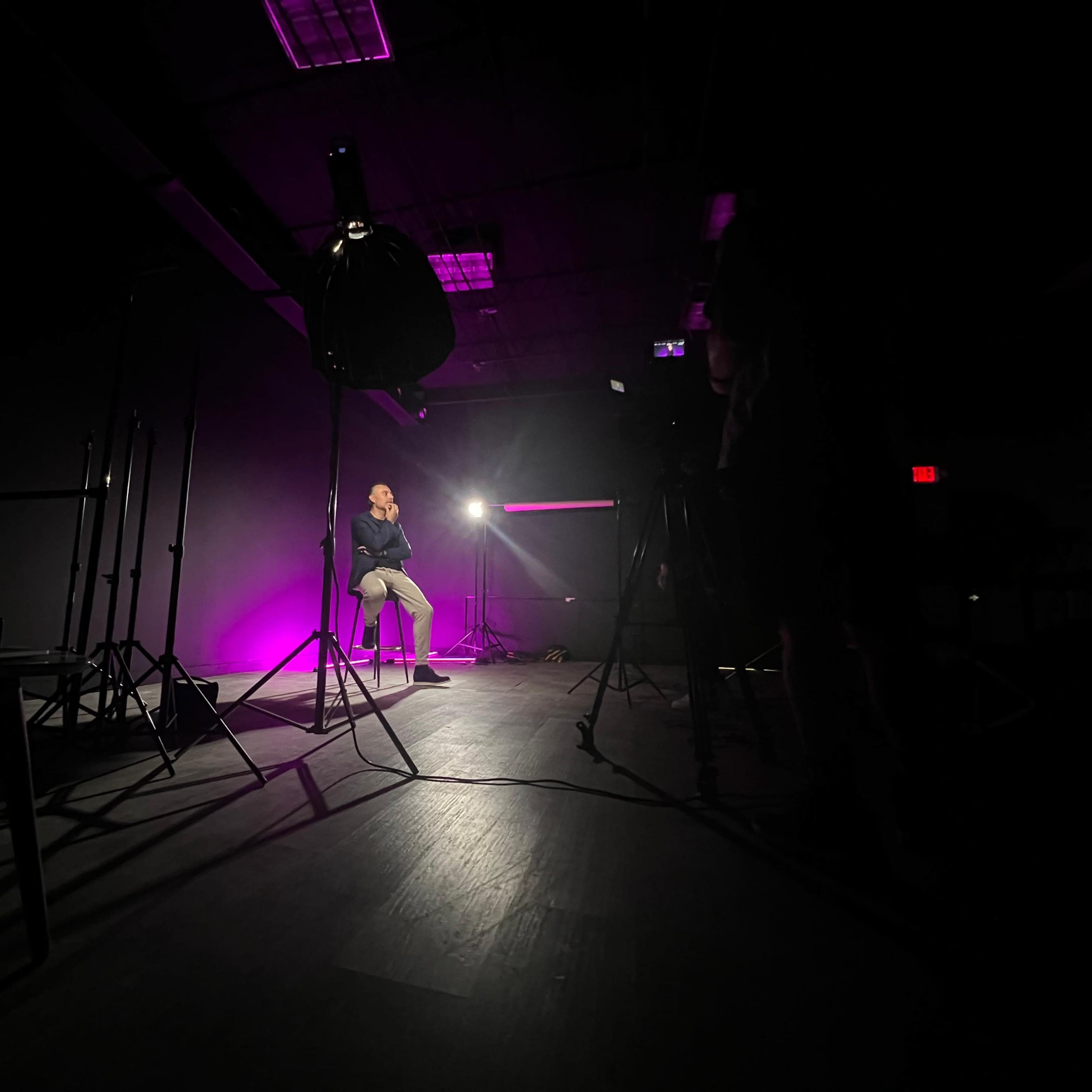 A person sits on a stool in a dimly lit studio, illuminated by a bright light with purple hues and surrounded by camera equipment and stands.