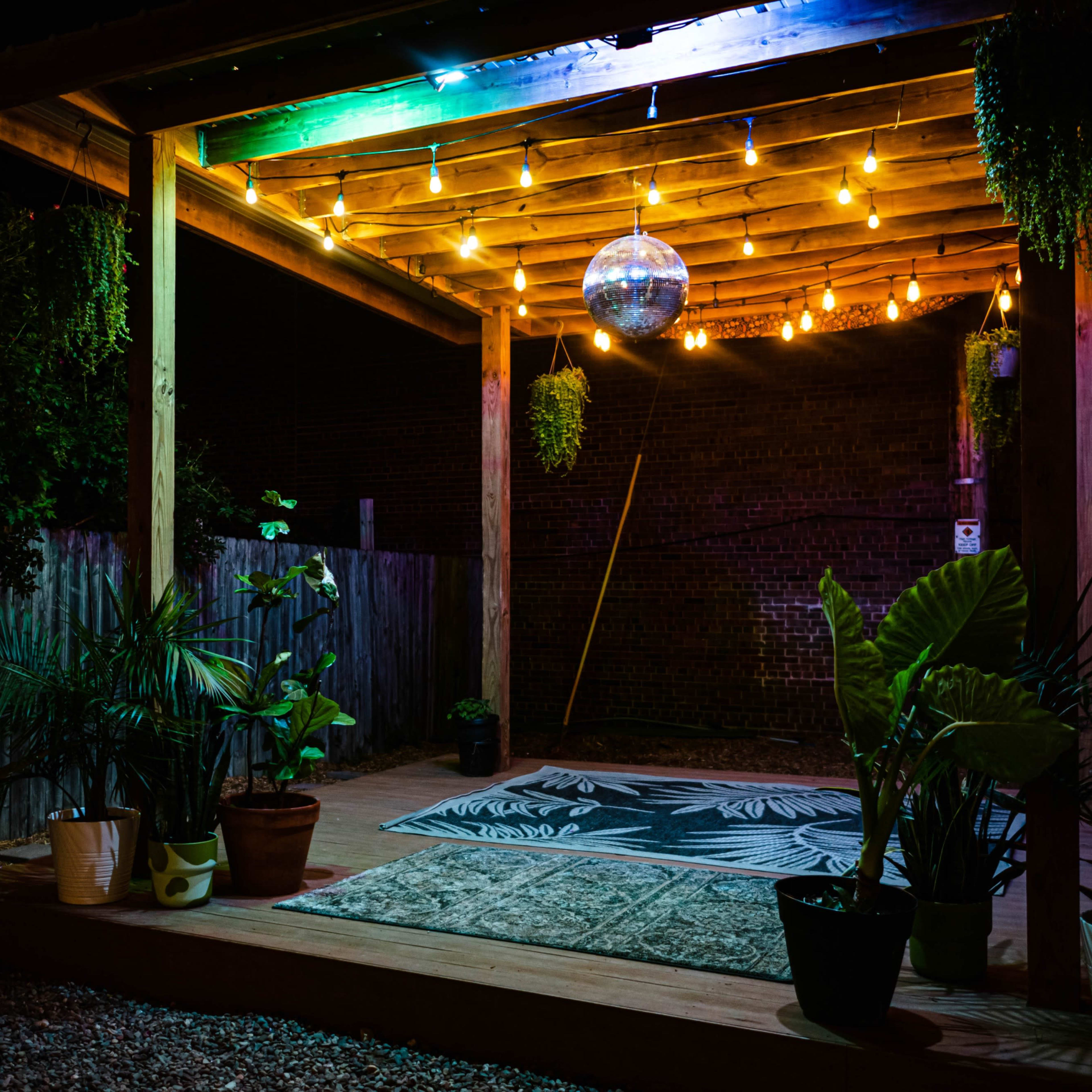 A wooden patio is illuminated by string lights and a disco ball, surrounded by potted plants and a patterned rug.