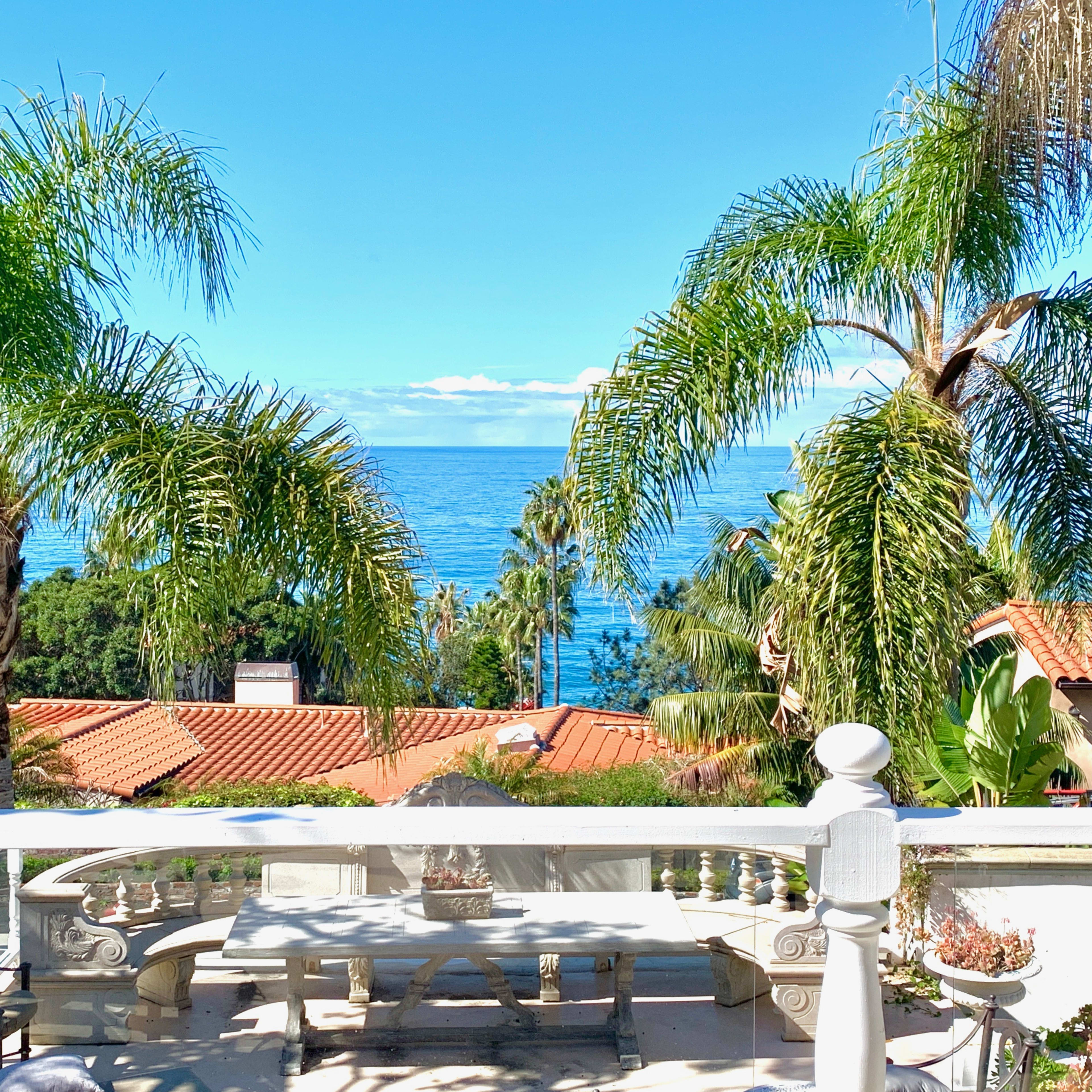 A balcony view shows a vibrant coastal landscape with palm trees framing the ocean and red-tiled roofs in the foreground.