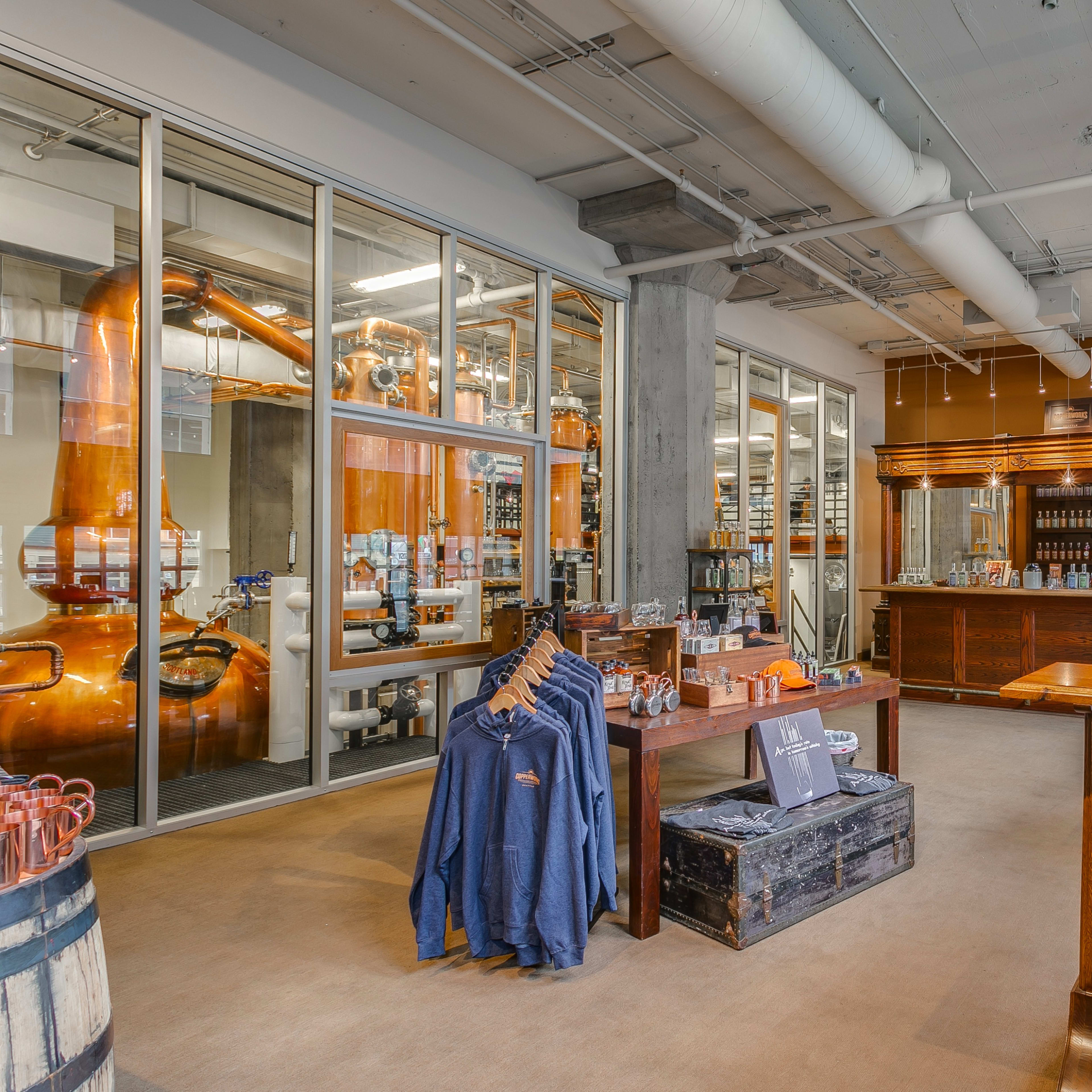 A spacious distillery bar area with large windows, a wooden counter, stools, and visible distillation equipment in the background.