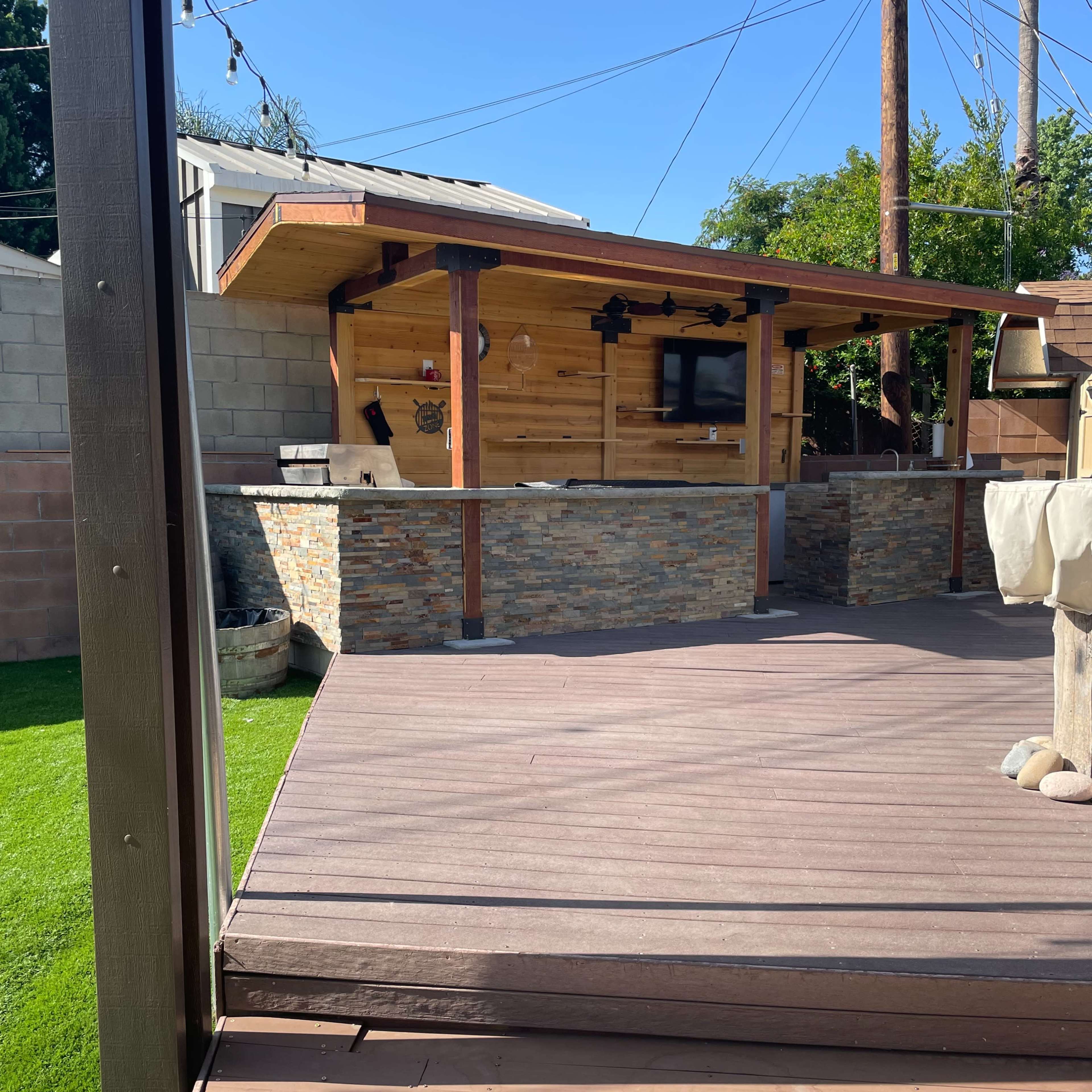 A wooden deck leading to an outdoor bar area made of stone and wood, surrounded by green grass and utility poles in the background.