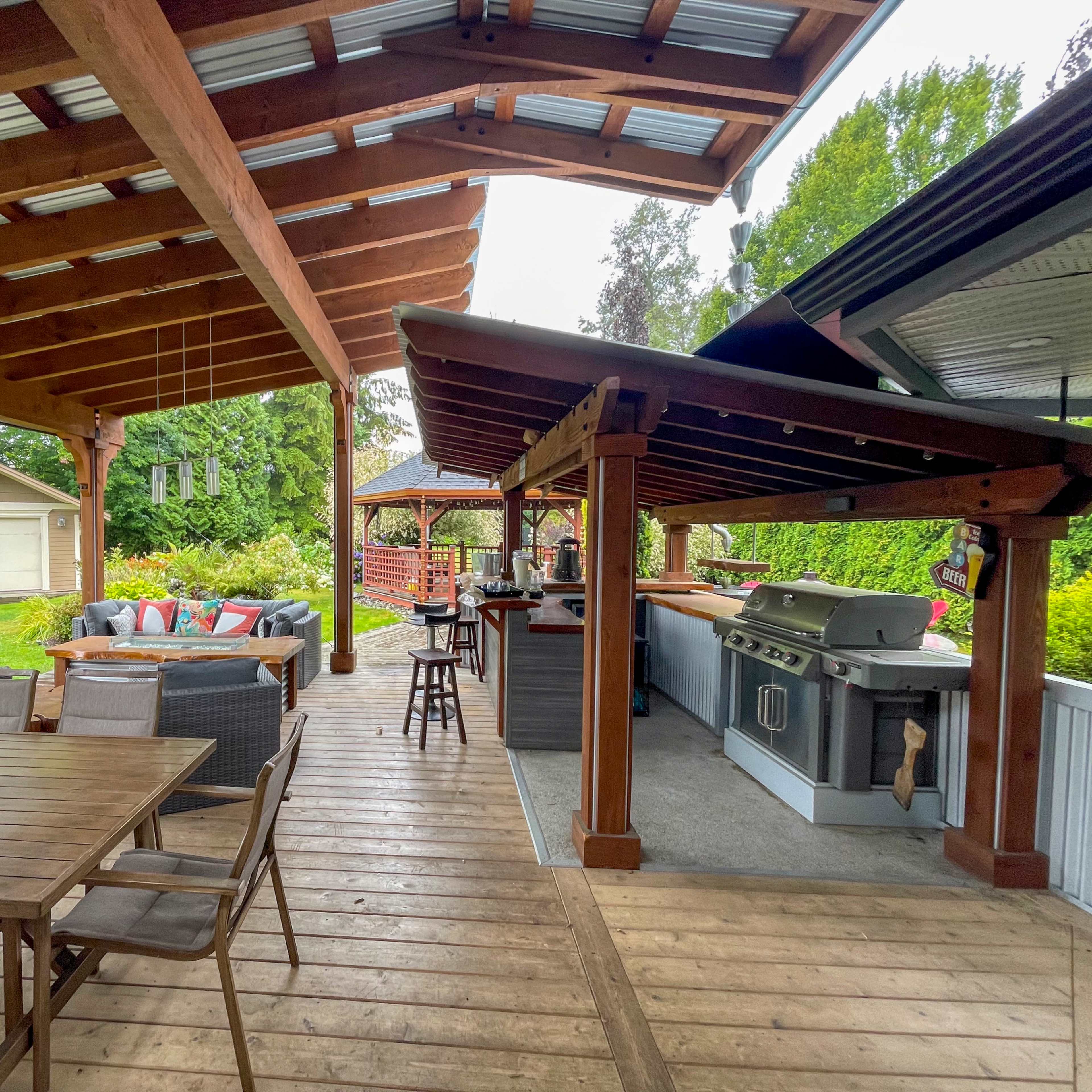A covered outdoor patio area with a dining table, lounge seating, and a barbecue grill setup, surrounded by greenery.