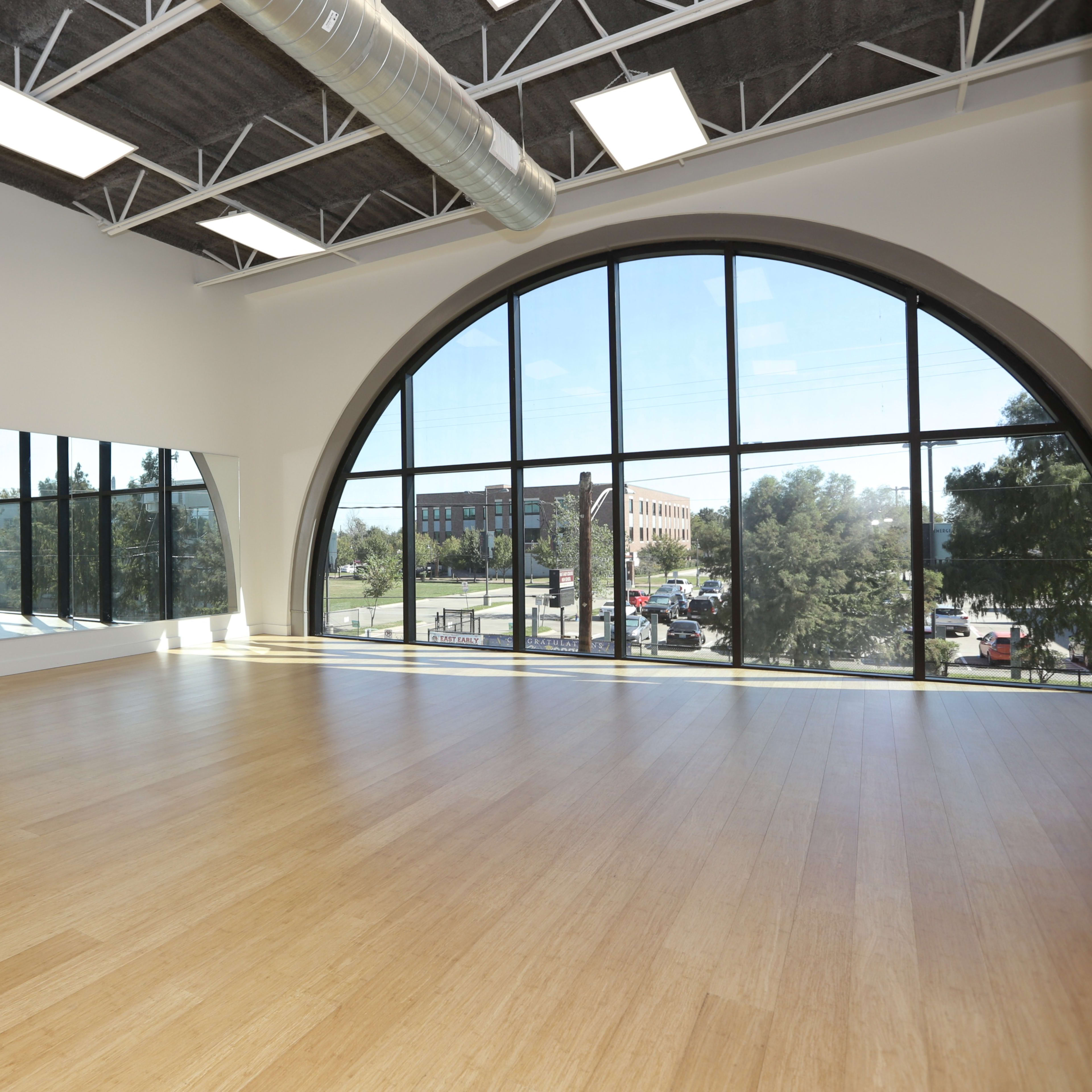 A large, well-lit dance studio features a curved window, a mirror along one wall, and a bamboo floor.