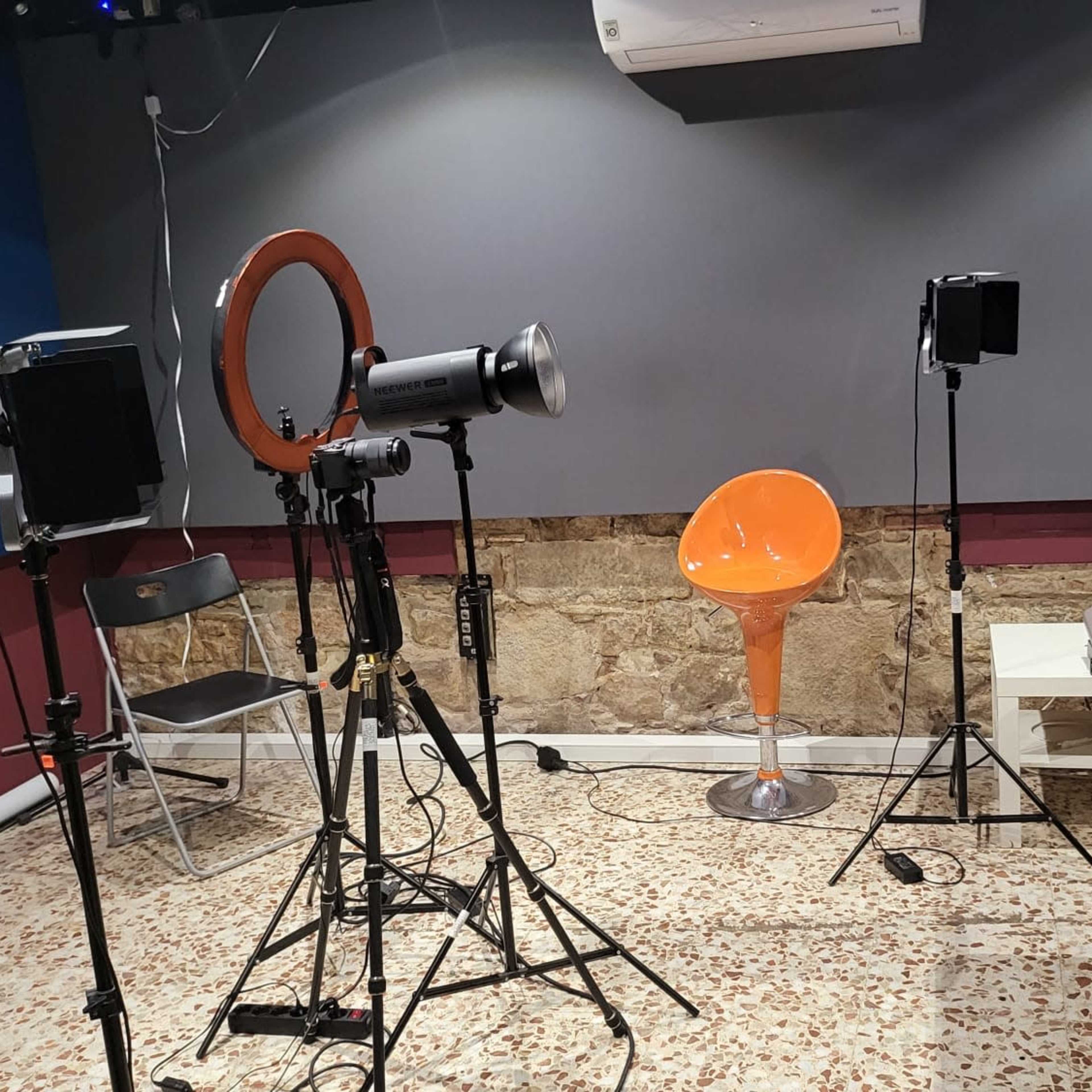 A studio setup with lighting equipment, an orange chair, a table with microphones, and a textured wall in the background.