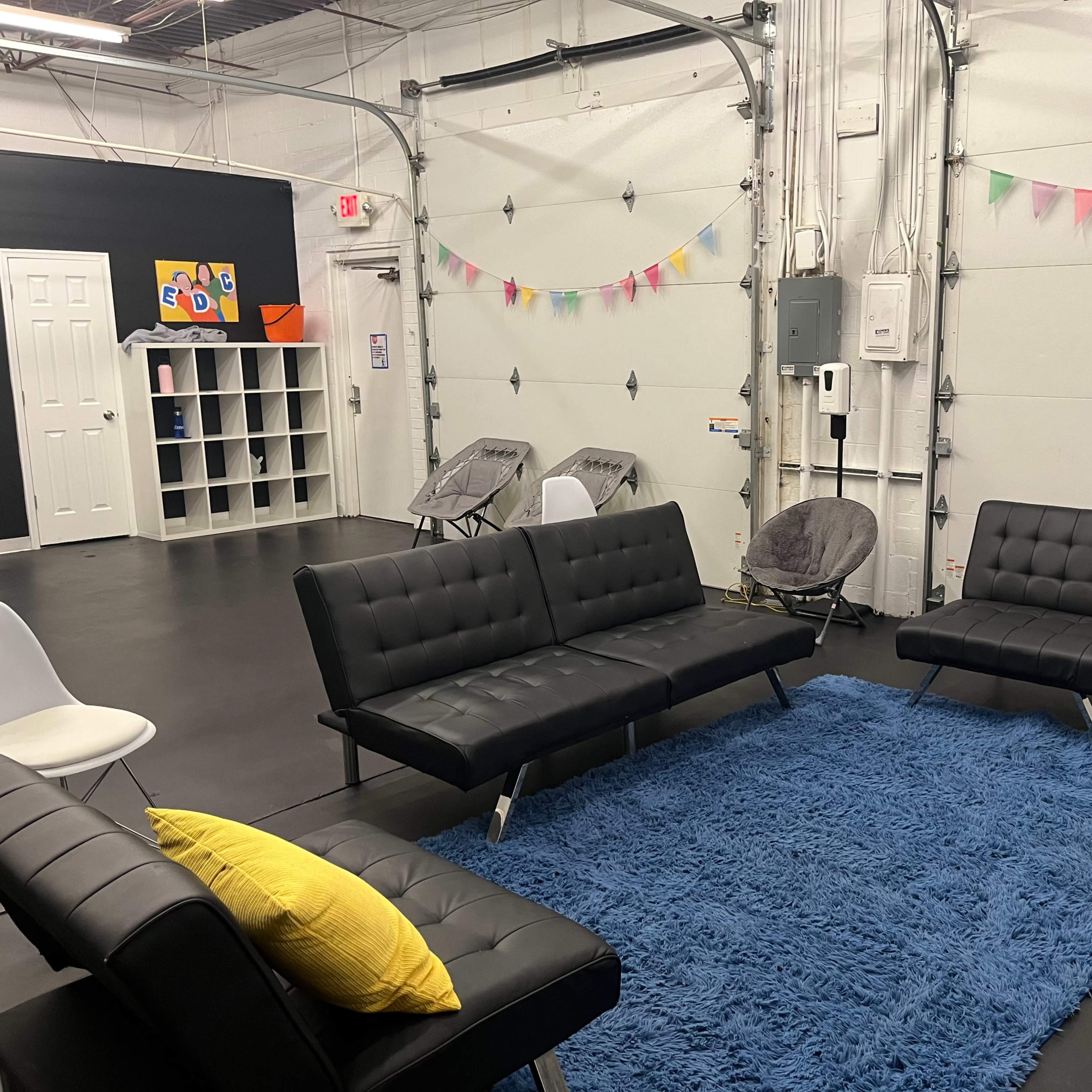 A modern, minimalistic lounge area with black sofas, a blue rug, and colorful decorations strung along the walls.