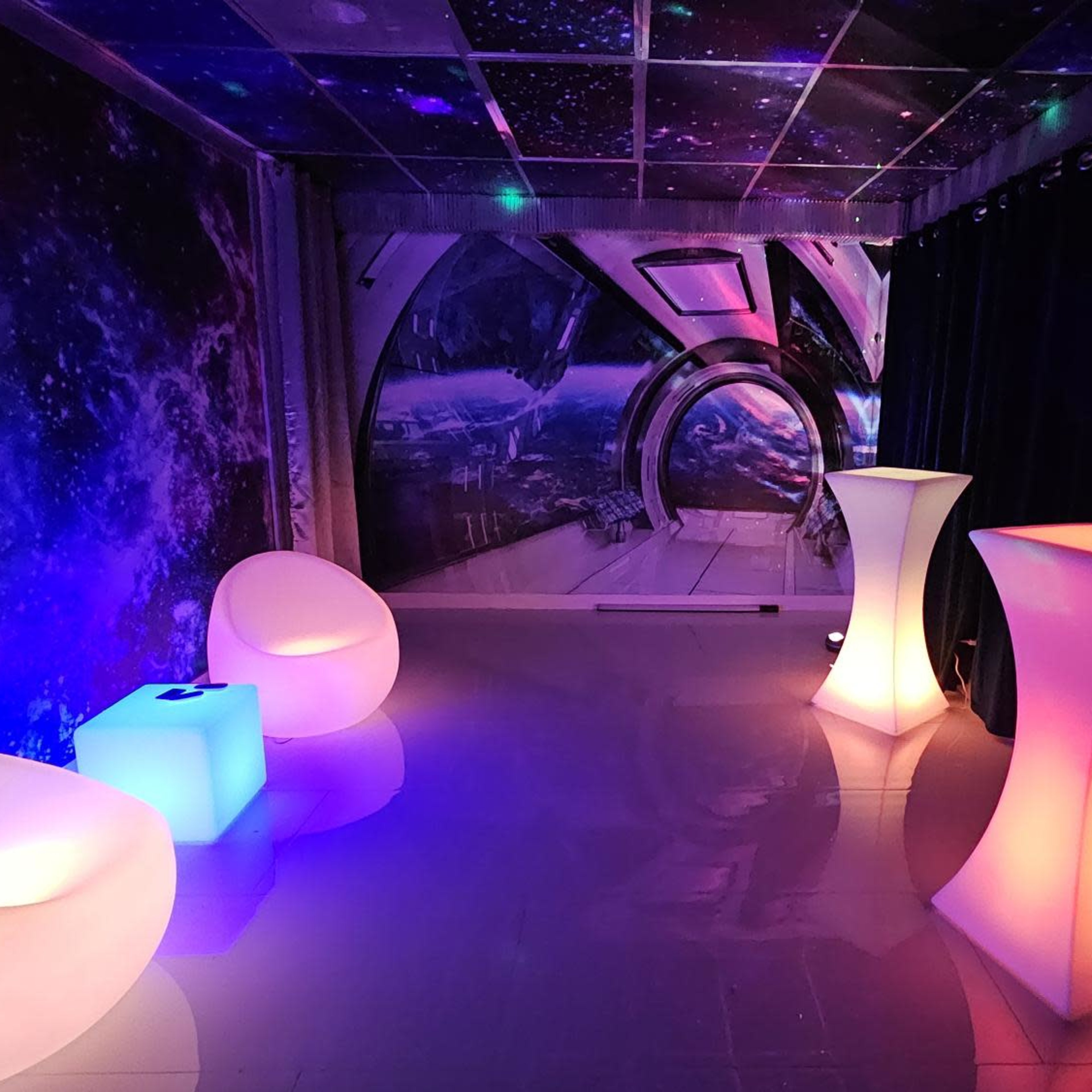 A room decorated with a cosmic mural and illuminated furniture, including two glowing tables and two curved chairs, set against dark curtains.