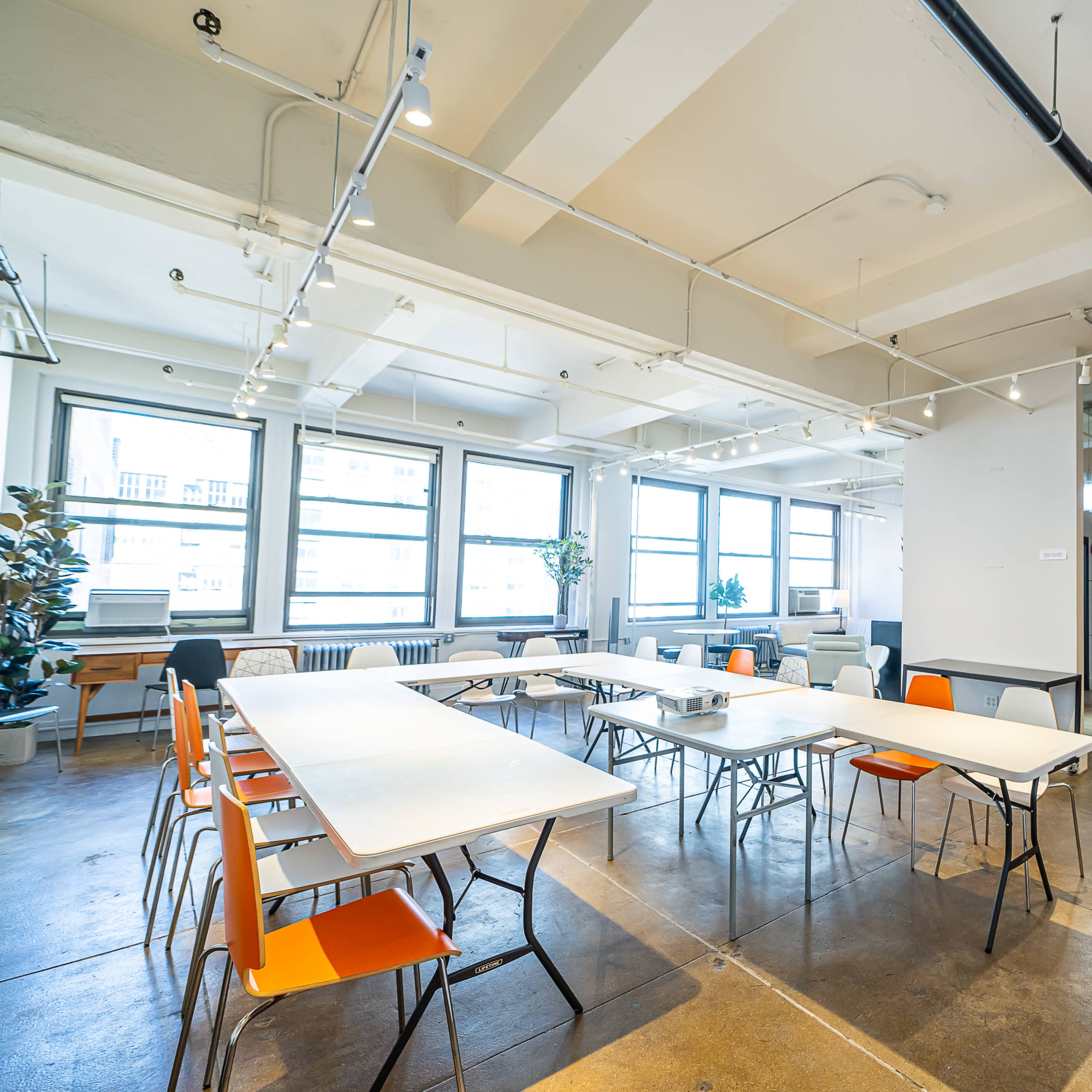 A bright, modern meeting room with several tables arranged in a U-shape, surrounded by chairs, large windows, and an accent wall in orange.