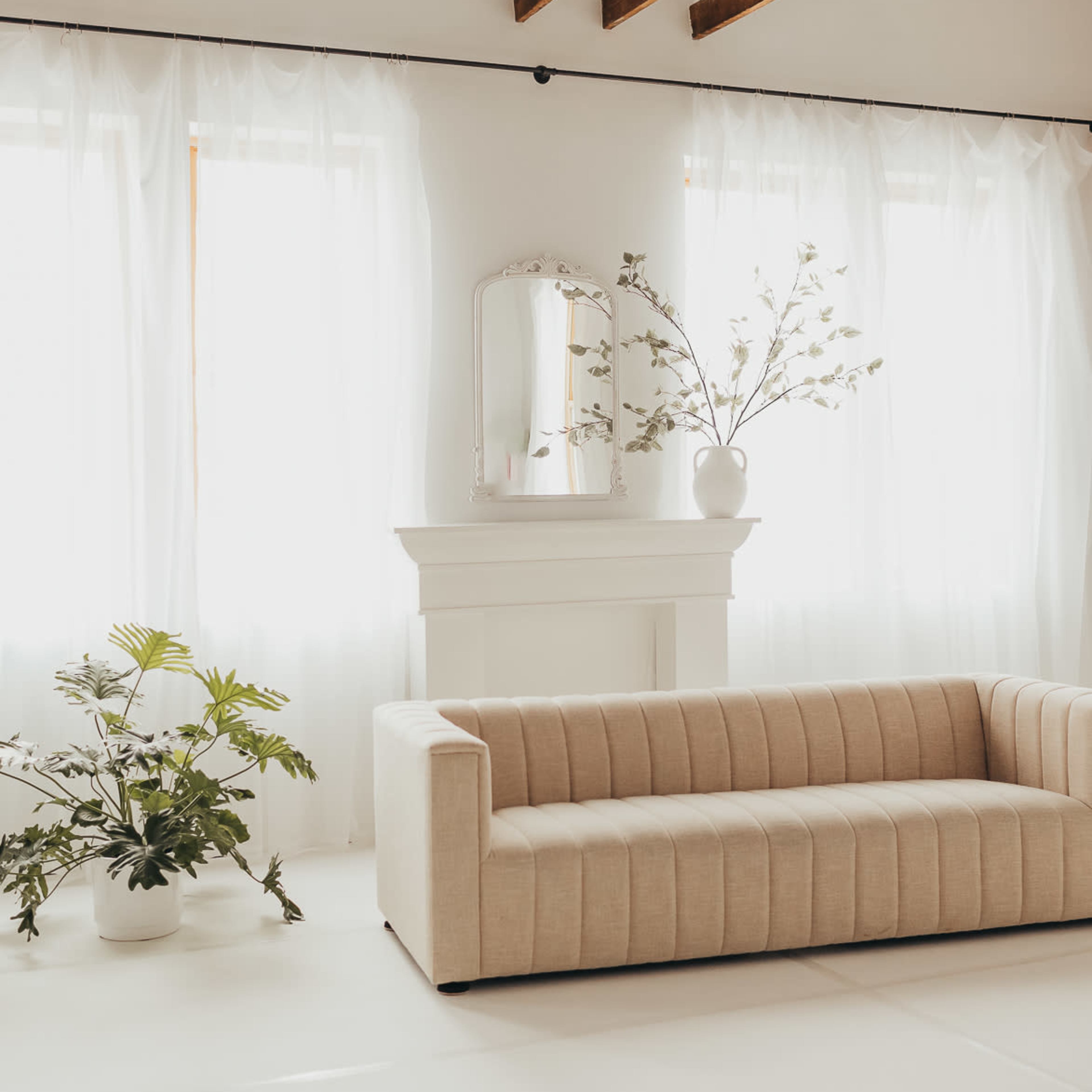 A beige, tufted sofa sits in a bright room with large windows draped in sheer curtains, beside potted plants and a decorative mirror on a white mantel.