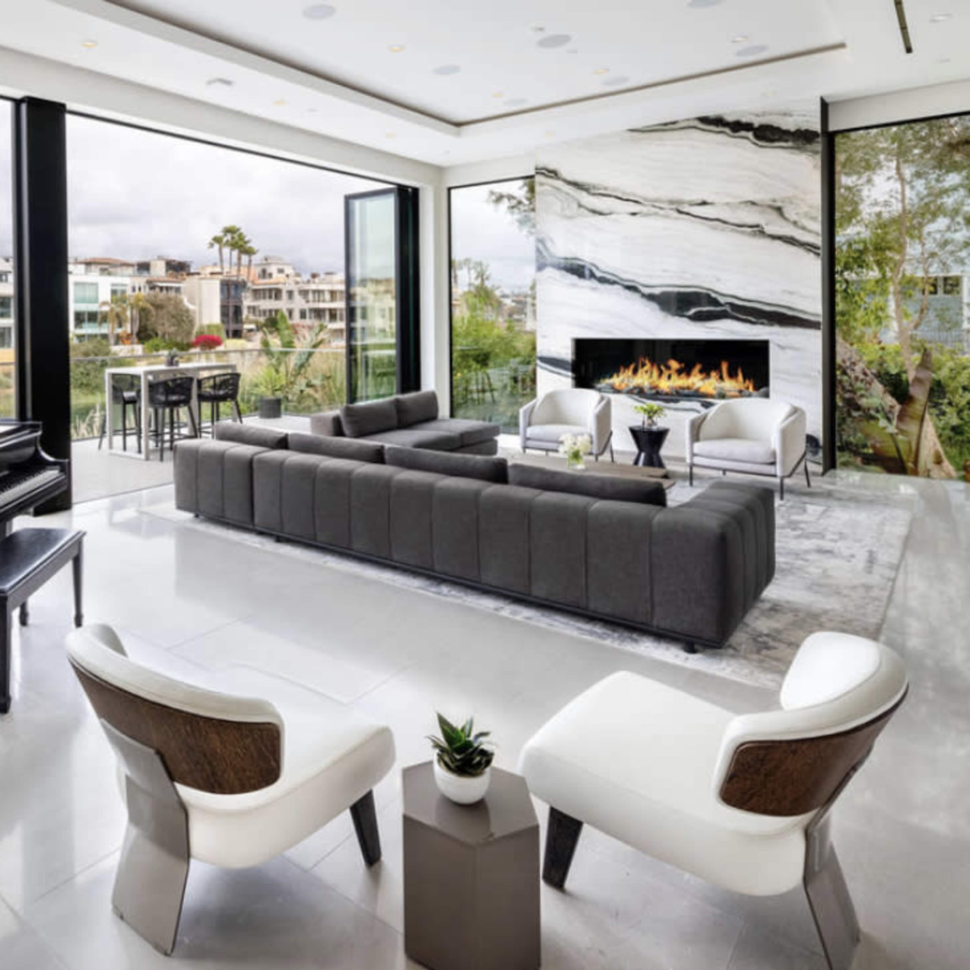 A modern living room featuring a grand piano, a sectional sofa, two white chairs, and large glass doors opening to an outdoor view with a fireplace and greenery.