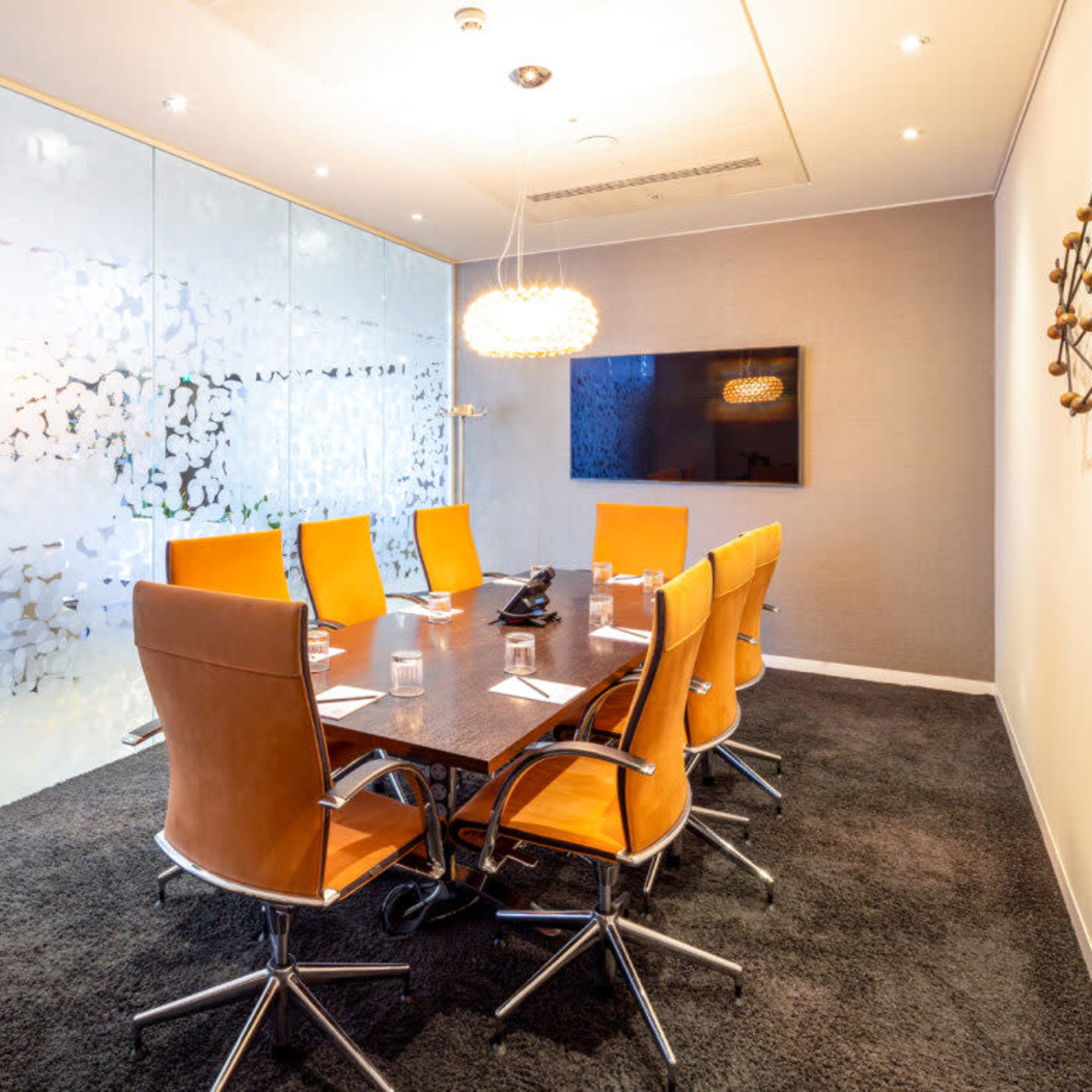 A modern conference room features a large table surrounded by orange chairs, with glass walls and a wall-mounted screen.