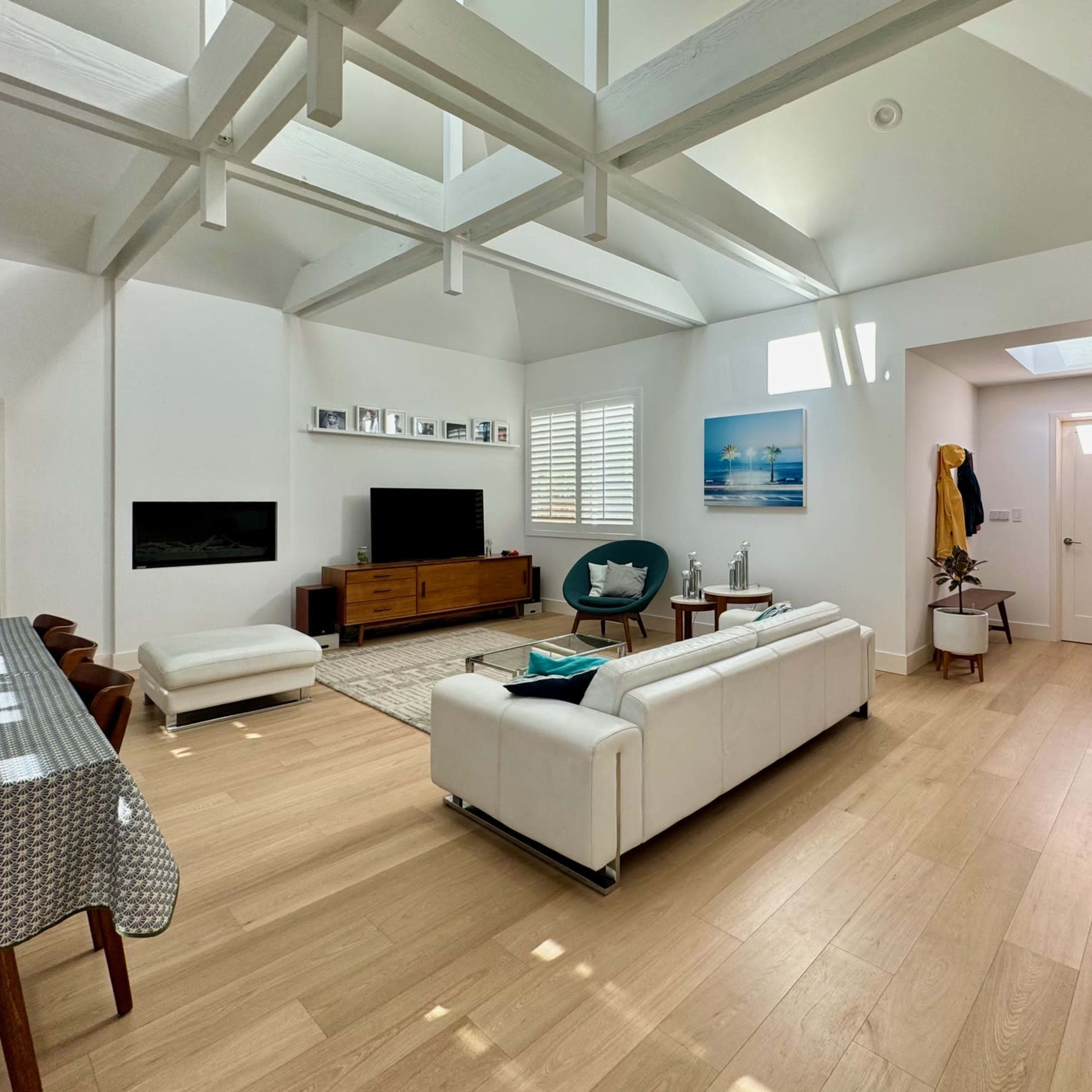 A bright, modern living room with a white couch, a wooden TV unit, a dining table, and large windows allowing natural light to fill the space.
