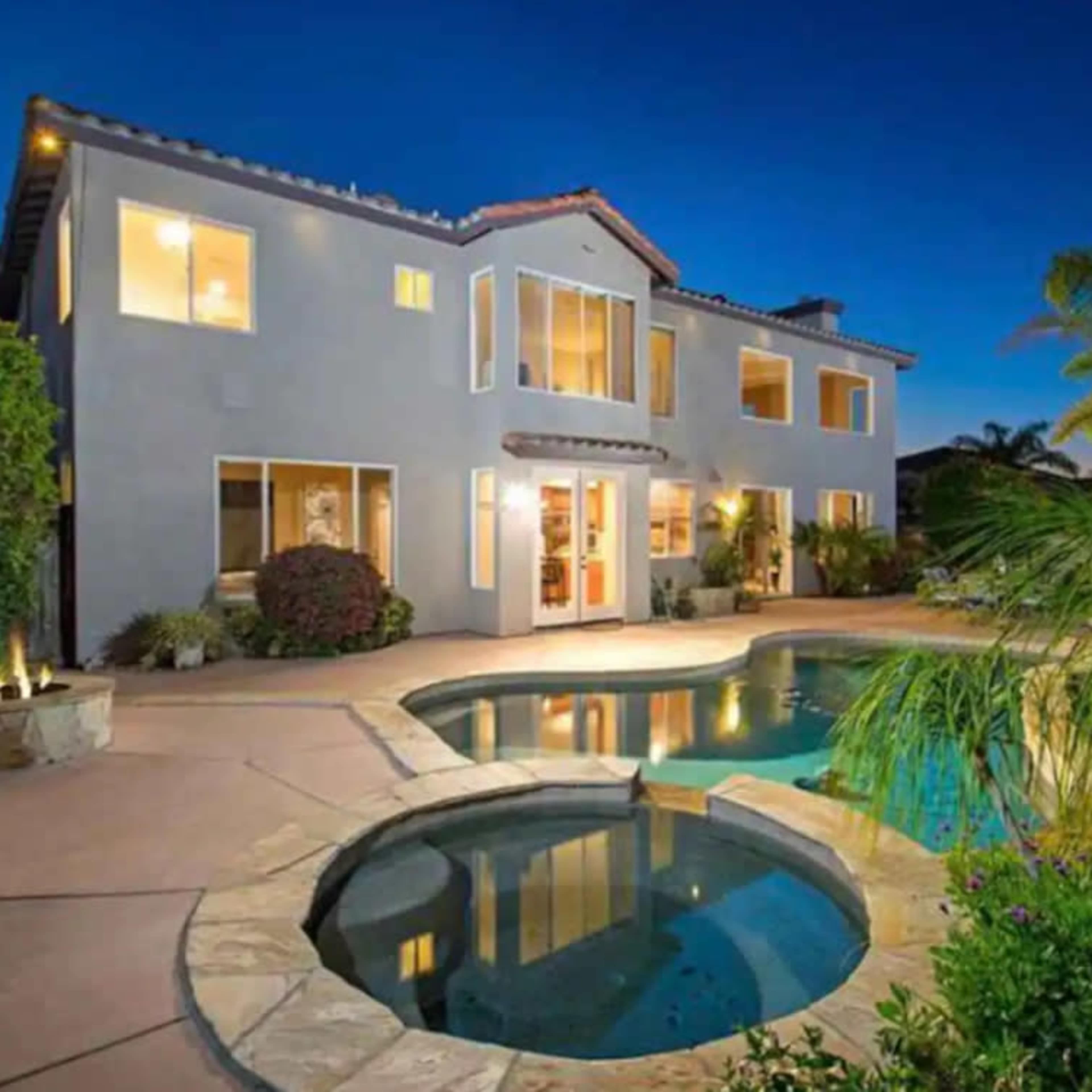 A two-story house with large windows and a swimming pool in the backyard, surrounded by palm trees and landscaping at dusk.