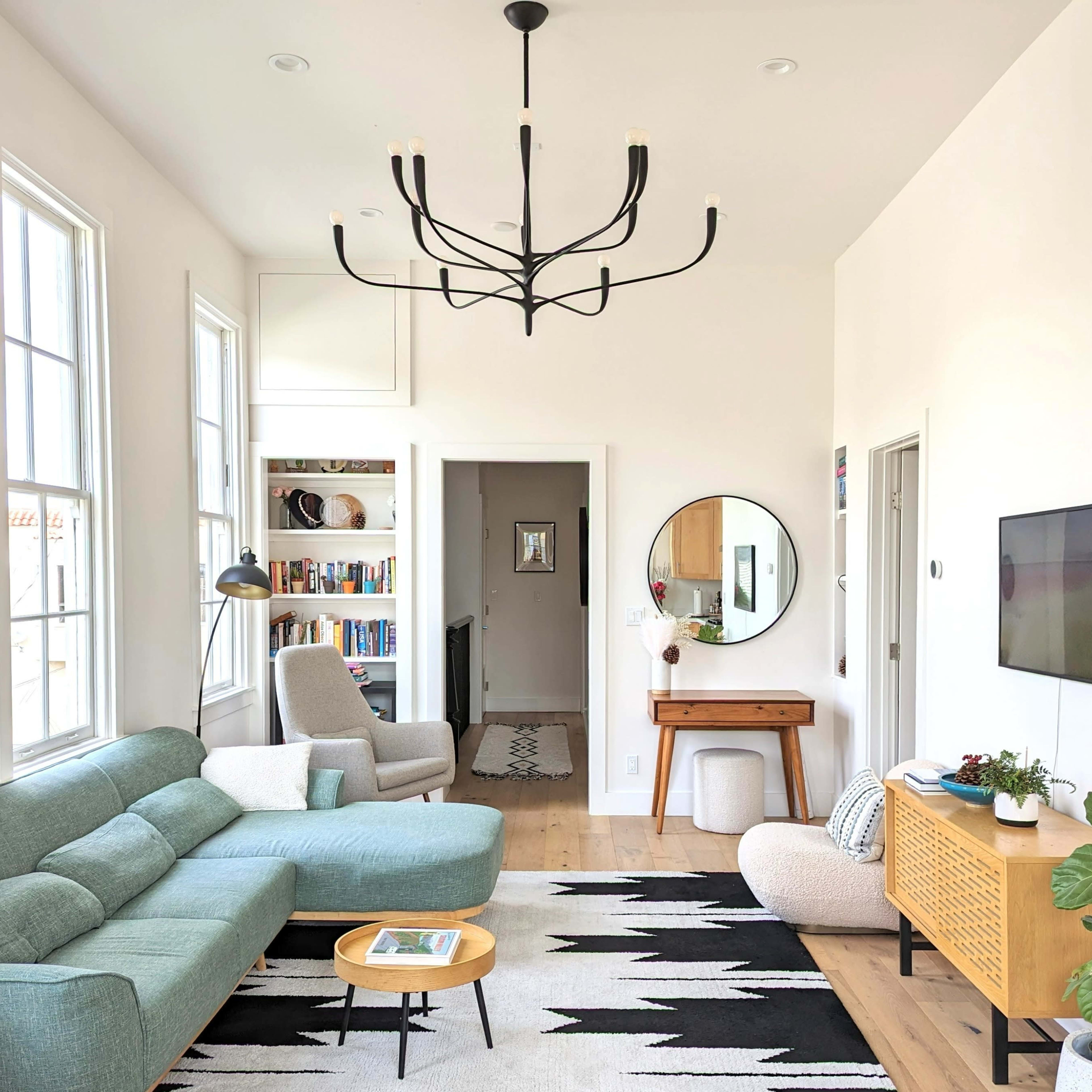 A modern living room with a green sofa, a round mirror, and a decorative chandelier, featuring a bookshelf and an entrance leading to another room.