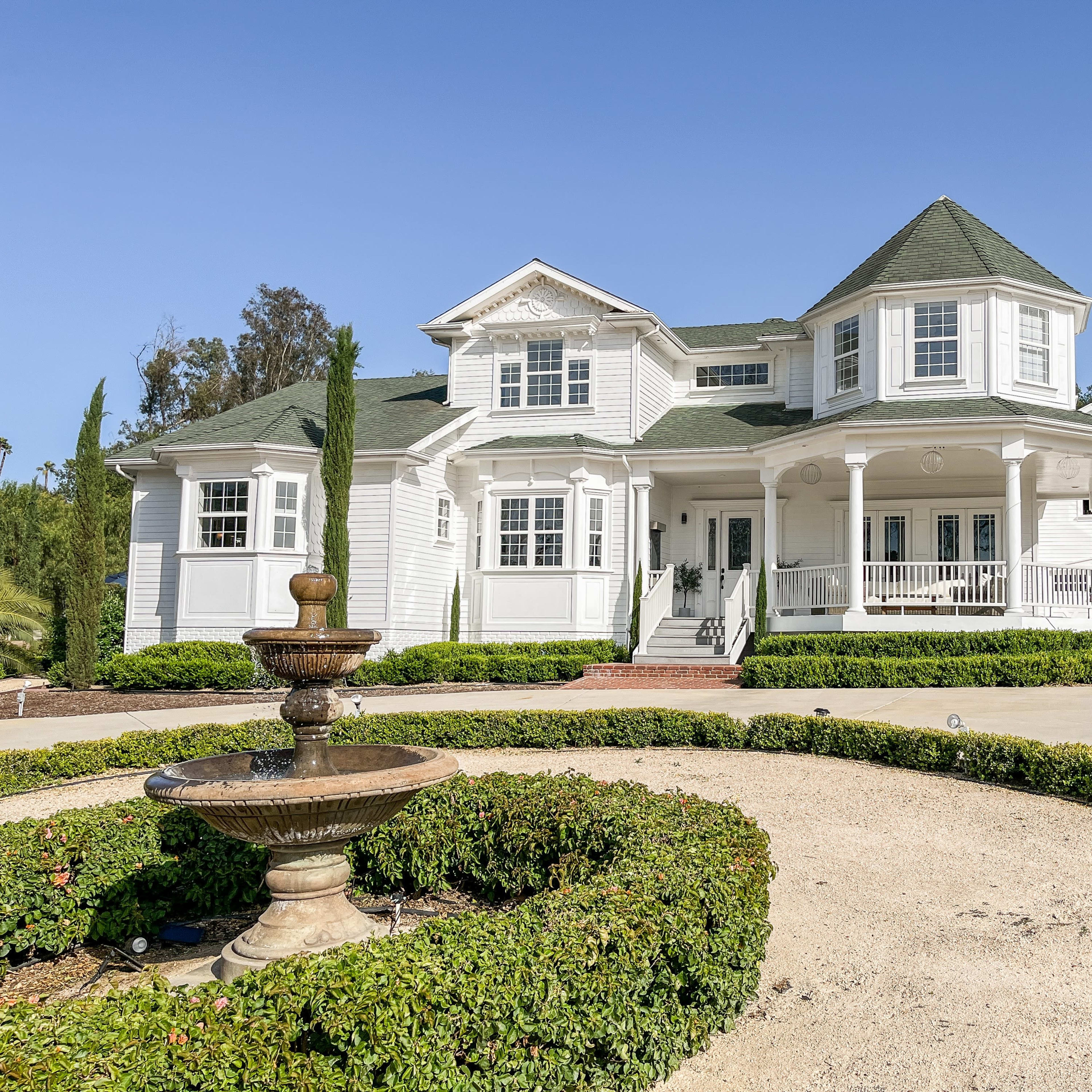 A large white house with a green roof, featuring a circular turret and a fountain in the front yard, surrounded by neatly trimmed hedges.