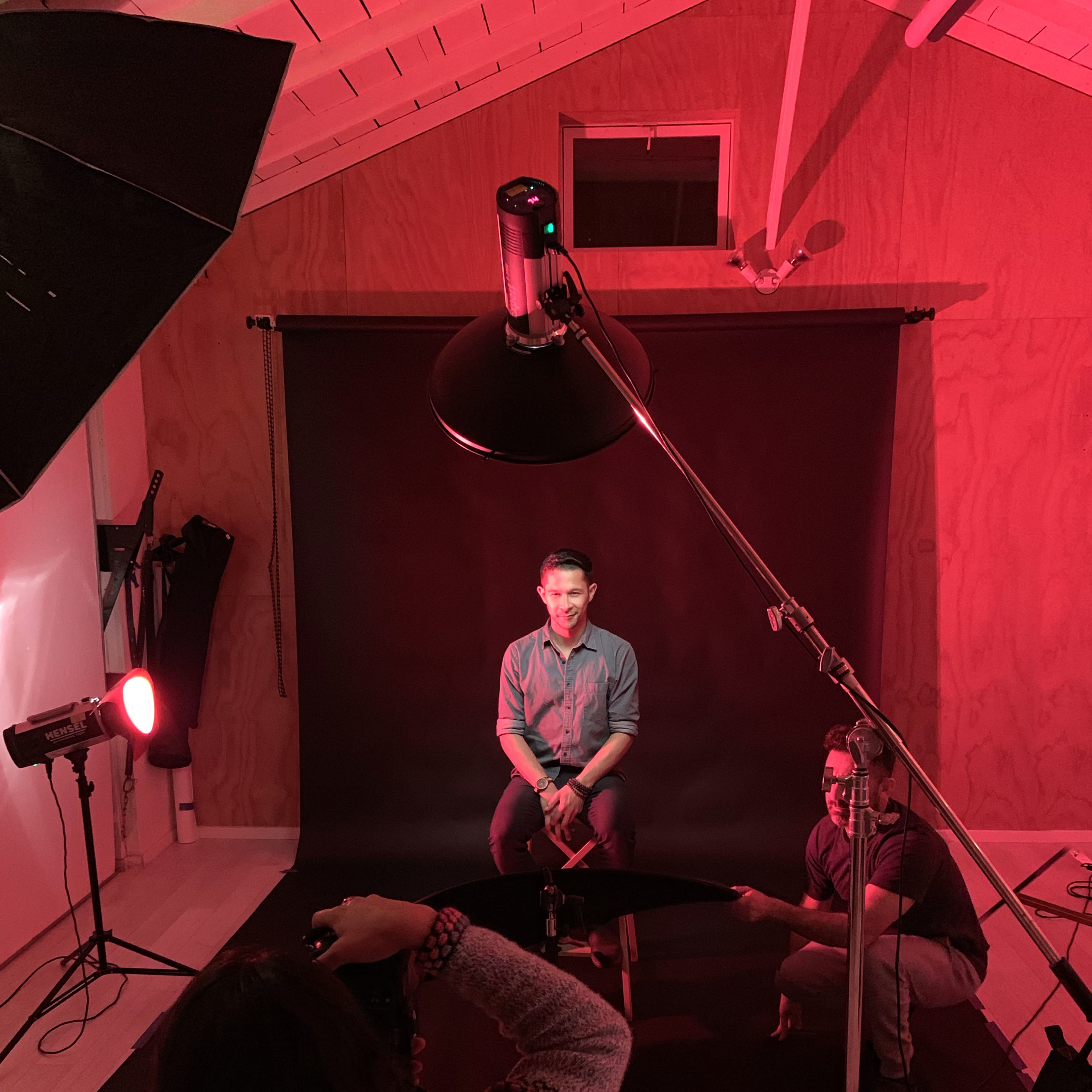 A person sits on a stool in front of a black backdrop while a photographer and an assistant work with lighting equipment in a studio setting.