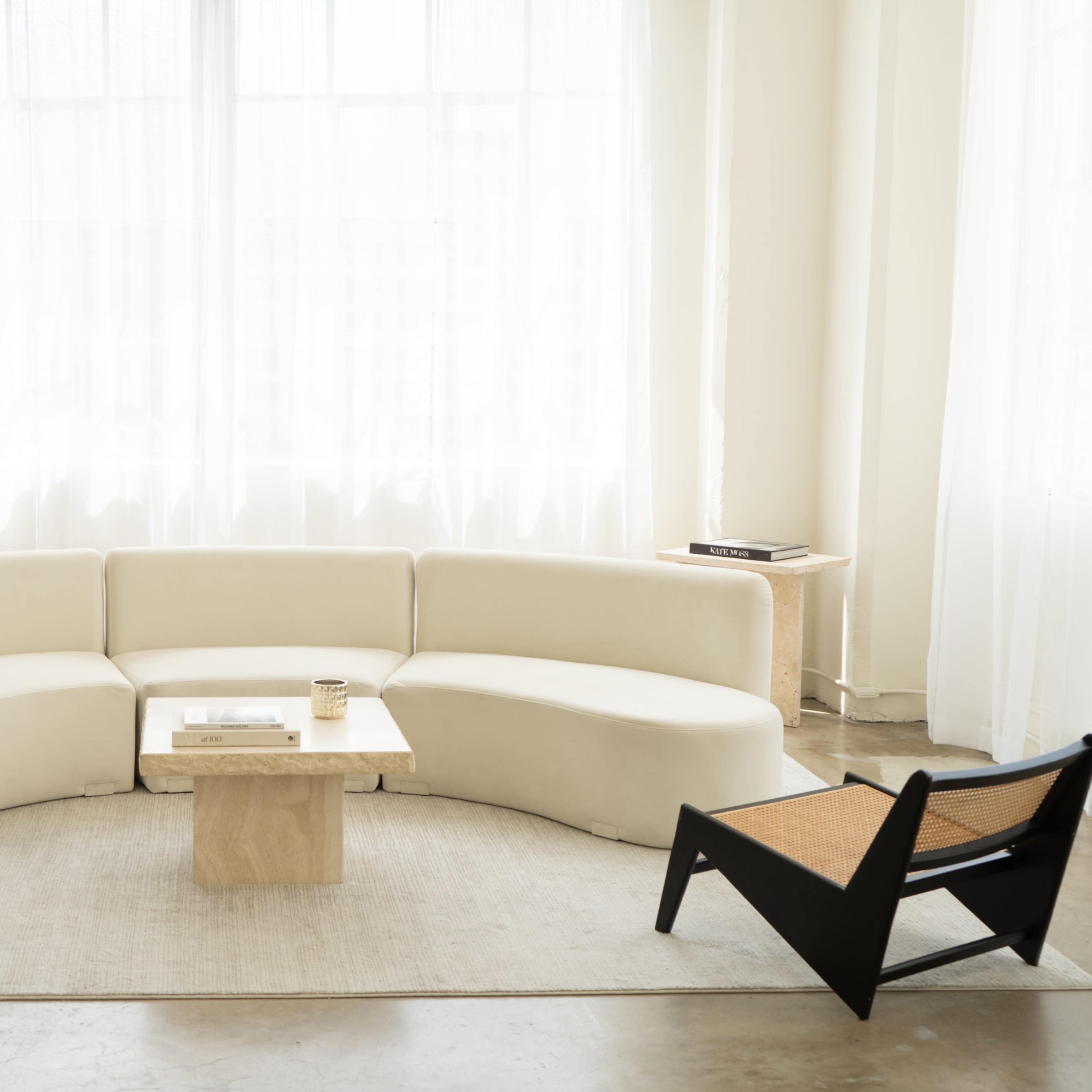 A minimalist living room with a curved white sofa, a wooden coffee table, and a single black chair, surrounded by large windows with sheer curtains.