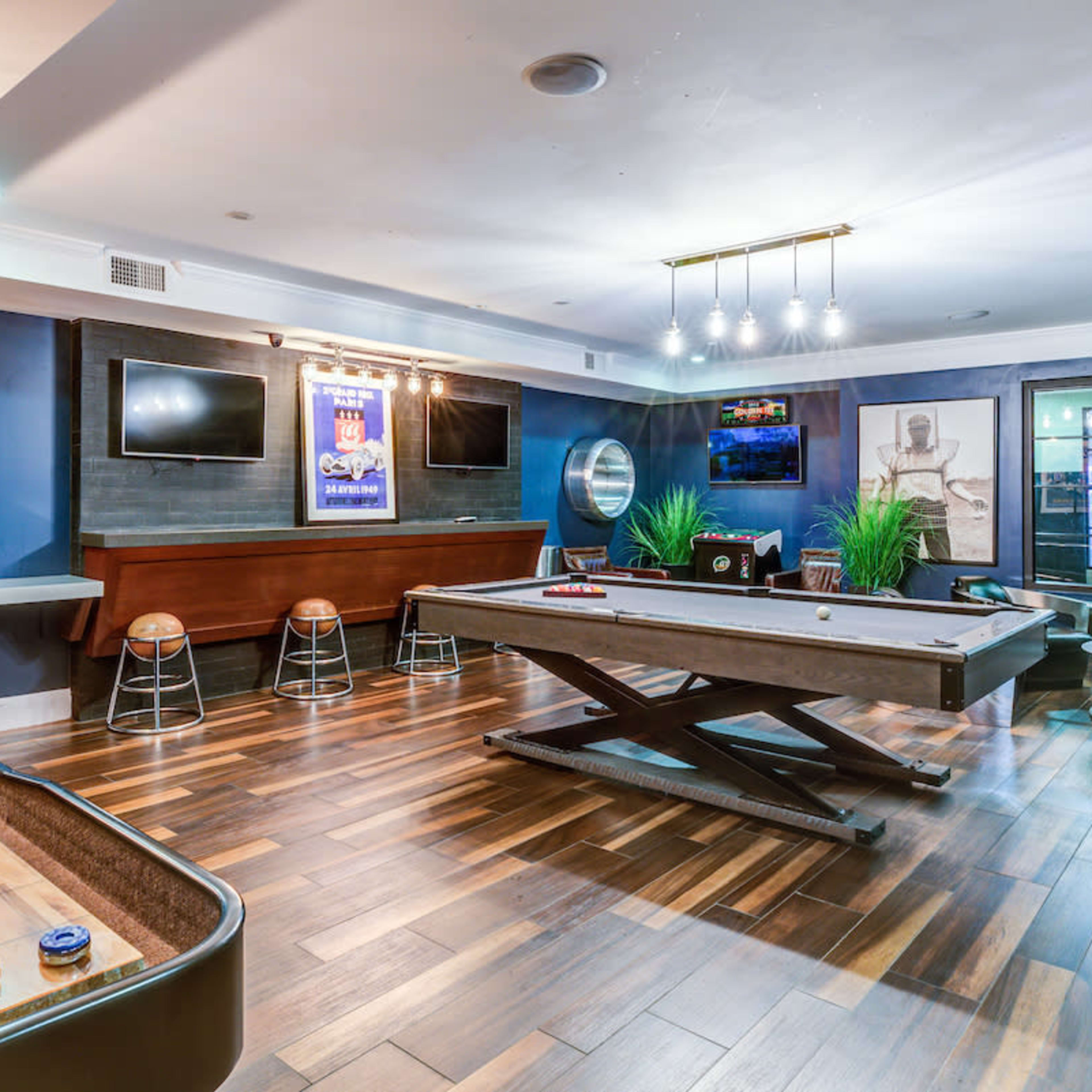 A recreational room featuring a pool table, a shuffleboard table, and several flat-screen TVs on the walls, with wooden flooring and modern lighting fixtures.