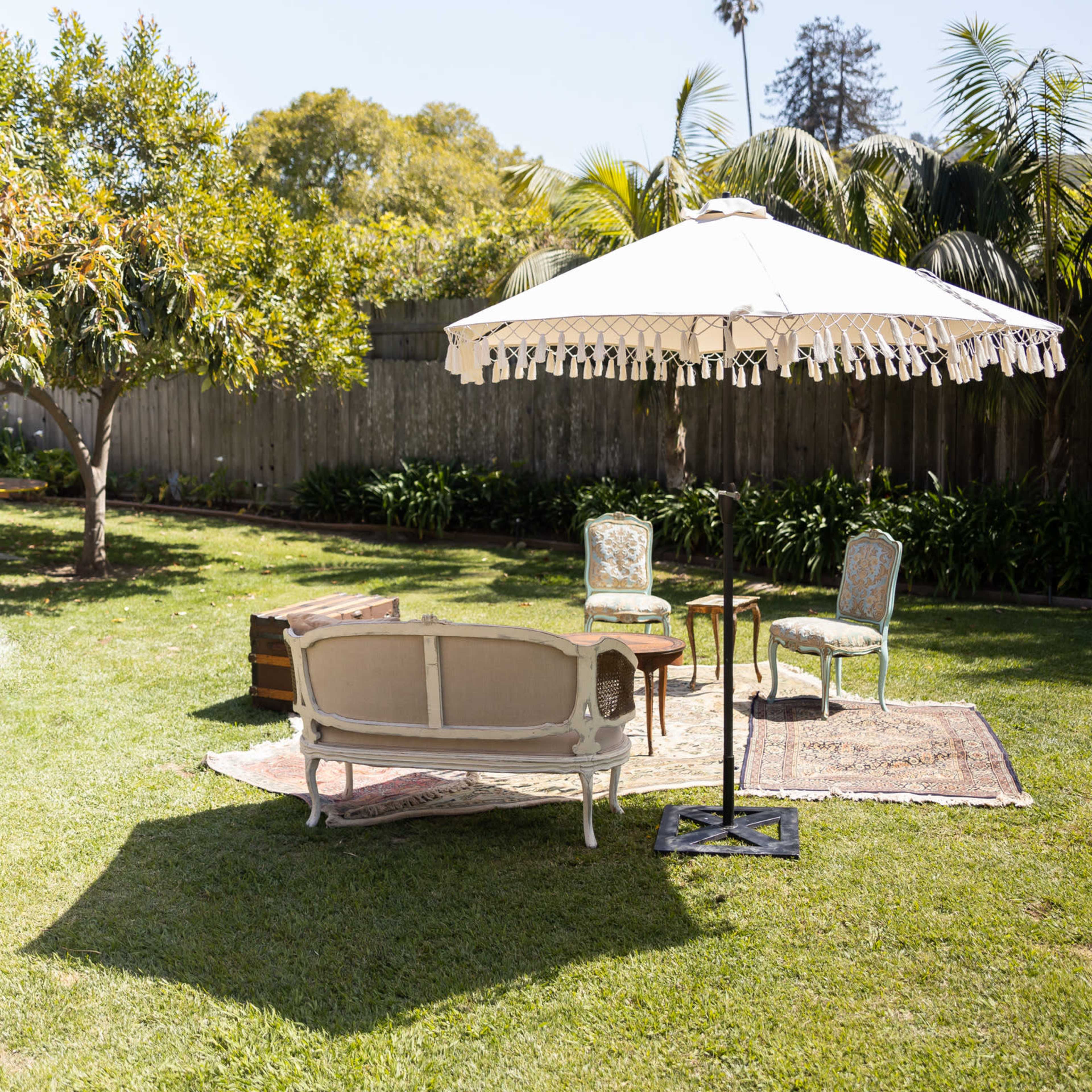 A sunlit backyard features a vintage couch, a small table, and a large umbrella on a patterned rug surrounded by greenery.