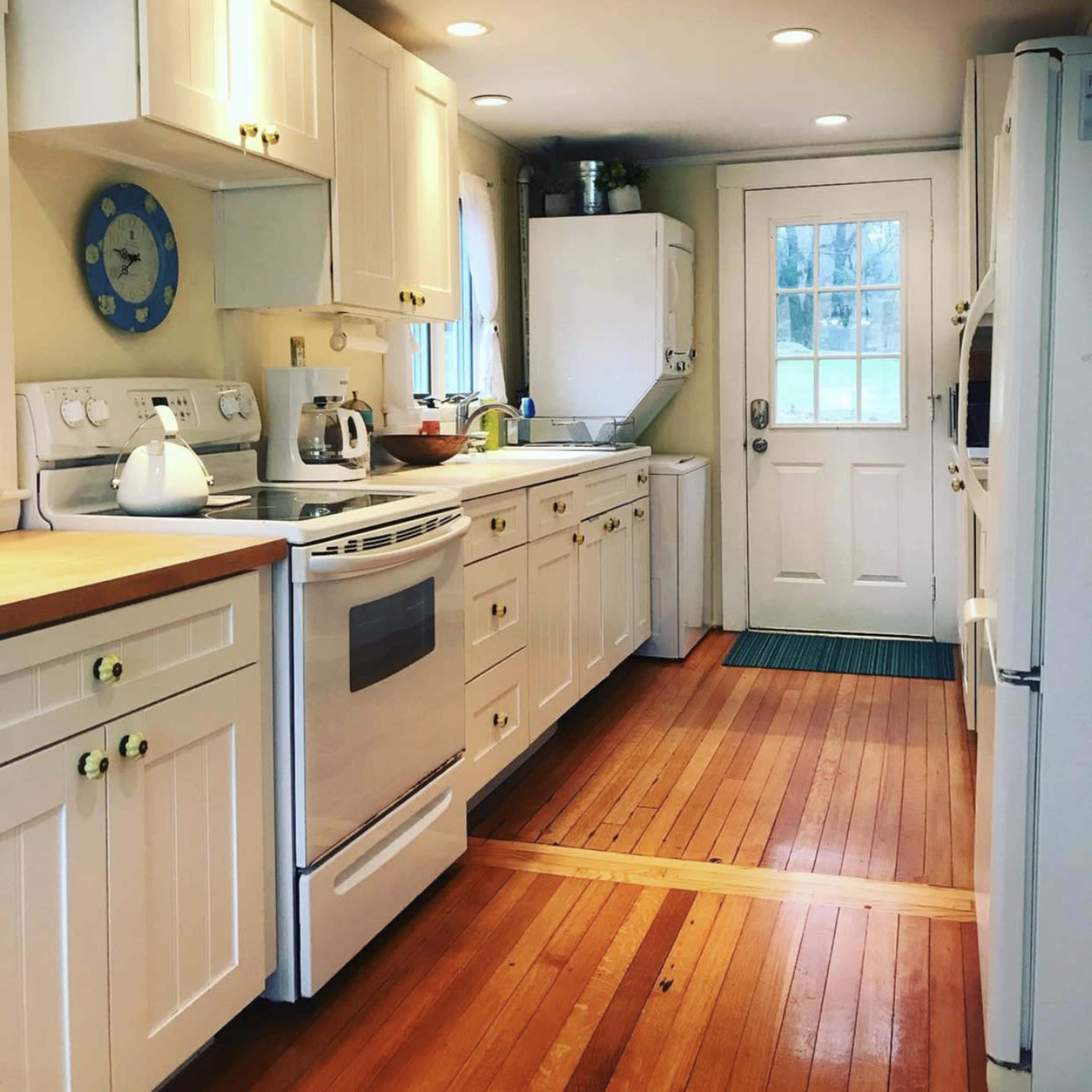 A narrow kitchen with white cabinets, wooden floors, and a door leading to the outside.