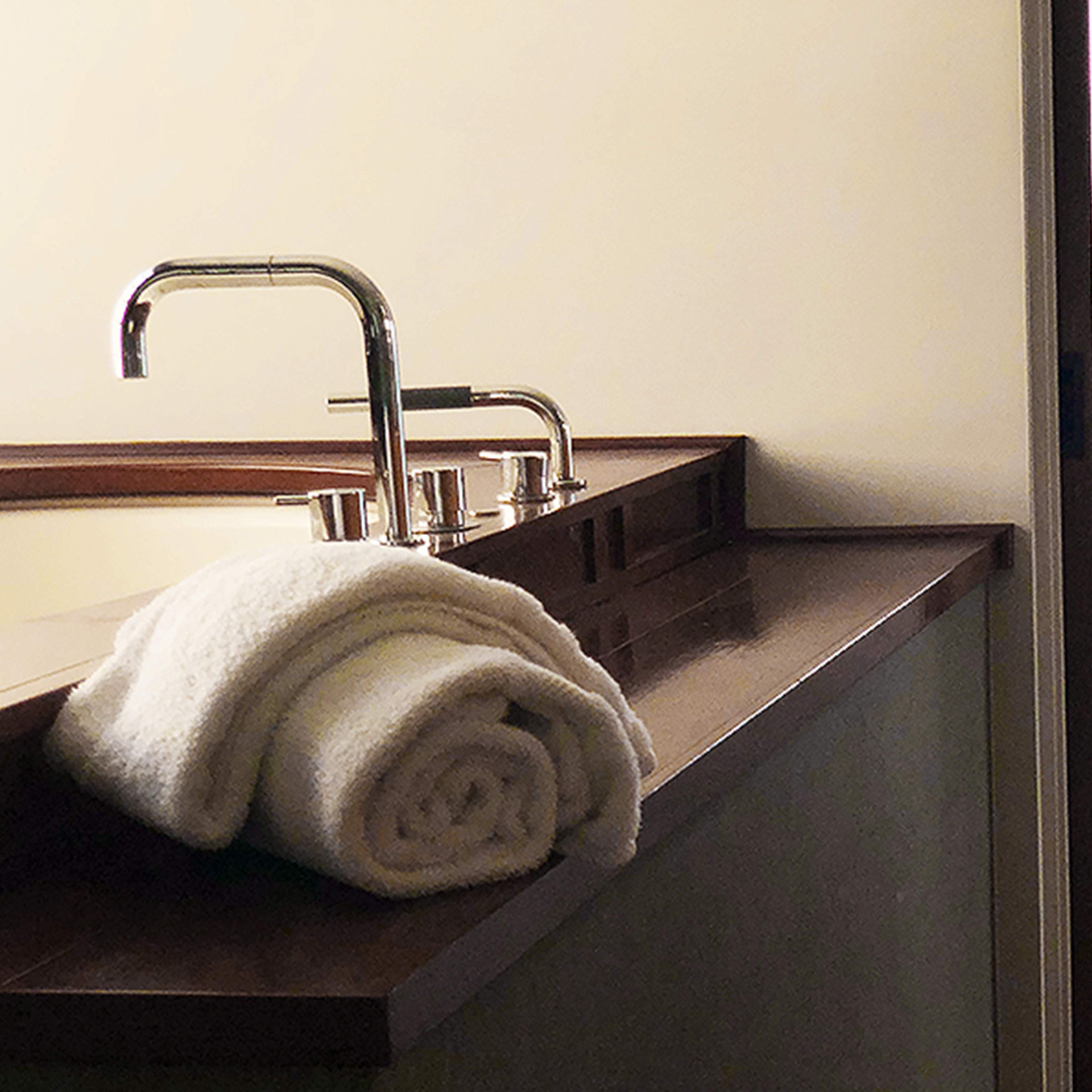 A bathroom area with rolled towels on a countertop, leading to a spacious room with a bed and a view of the ocean through an open door.