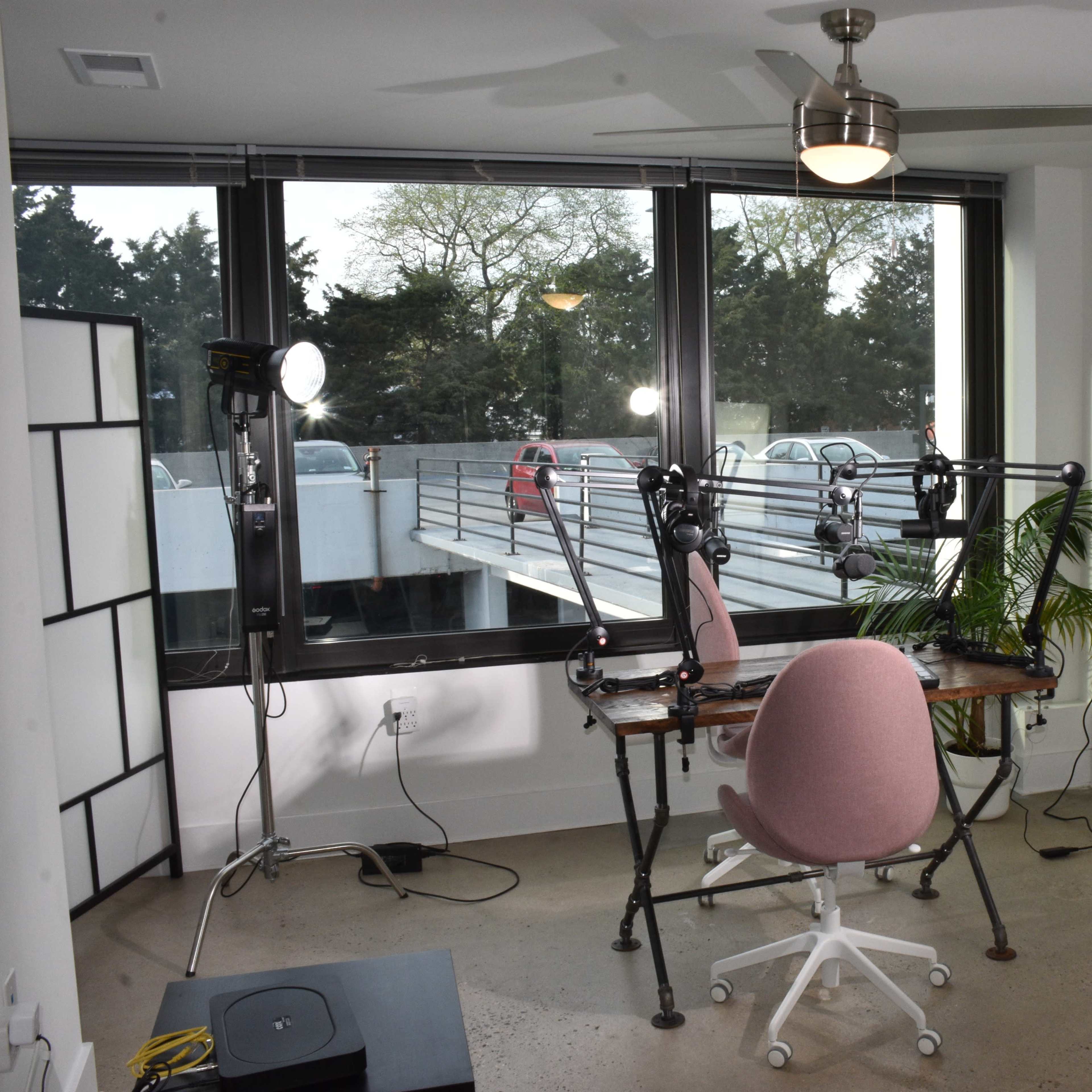 A modern office space with a desk, a pink office chair, and two bright lights set up for video recordings, with large windows providing an outside view.