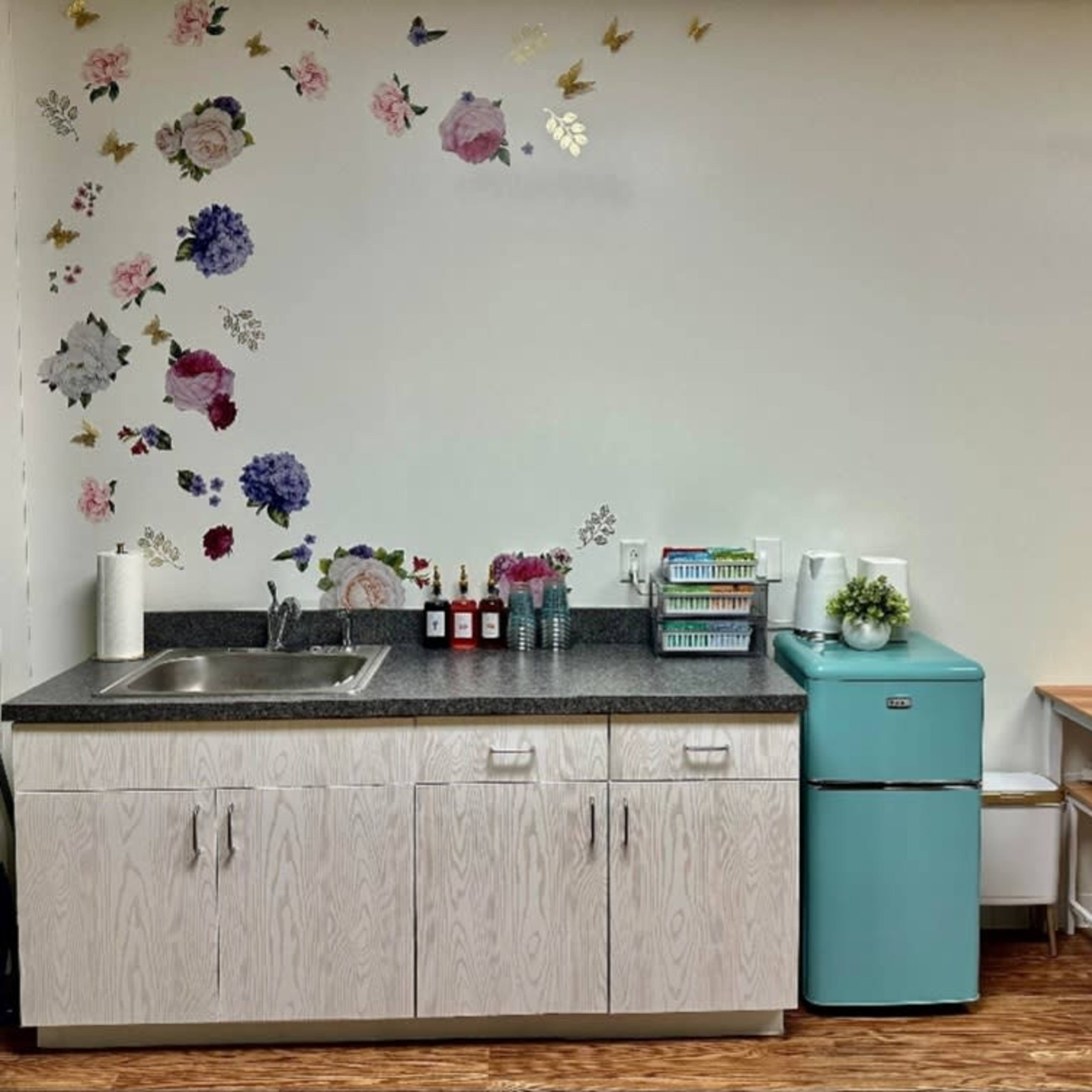 A small kitchen area features a sink, a turquoise fridge, and a wooden table, with floral decorations on the wall.