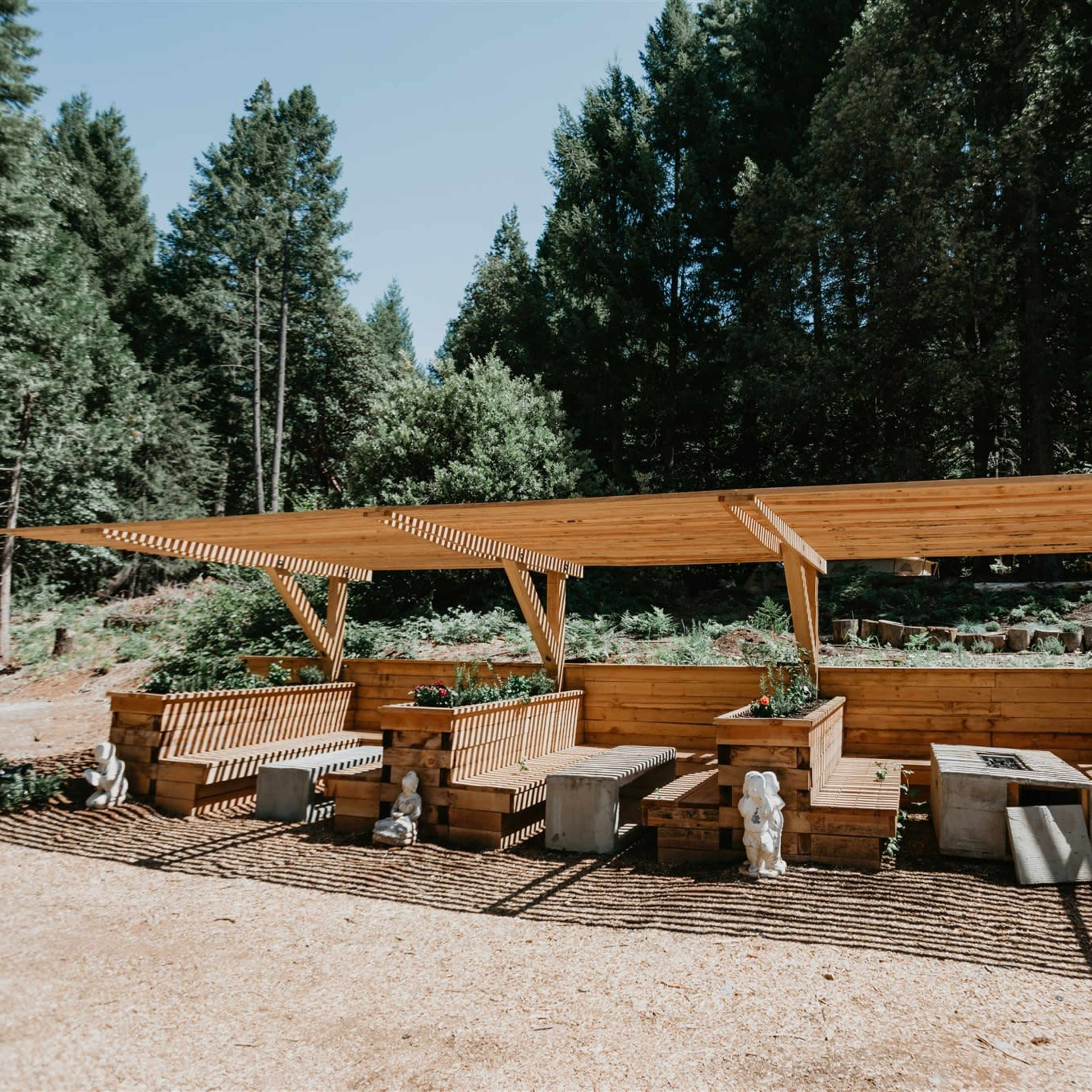 A wooden outdoor seating area with planters and a shaded roof, surrounded by trees.