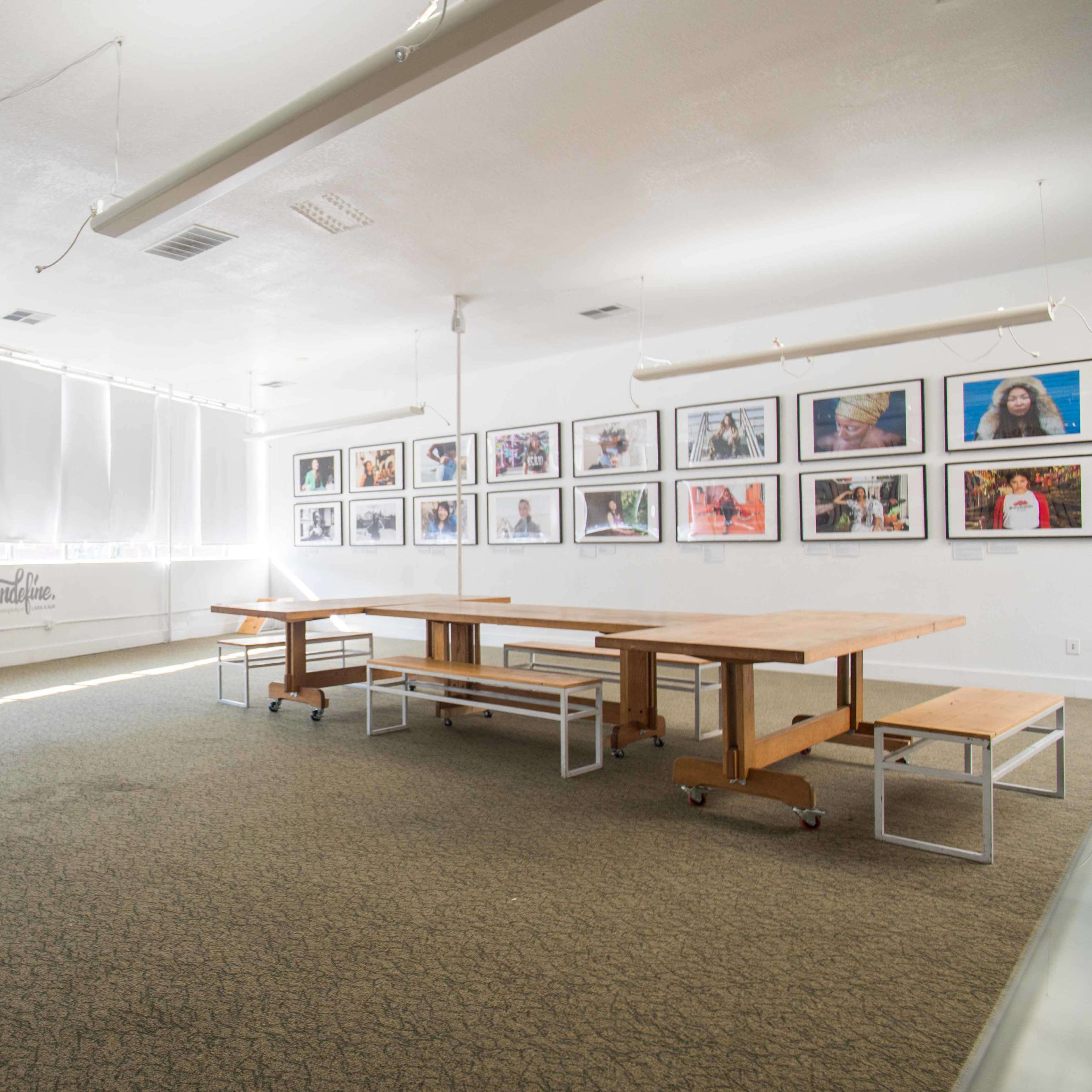 A spacious, well-lit room with large windows, featuring several tables and a wall adorned with framed photographs.