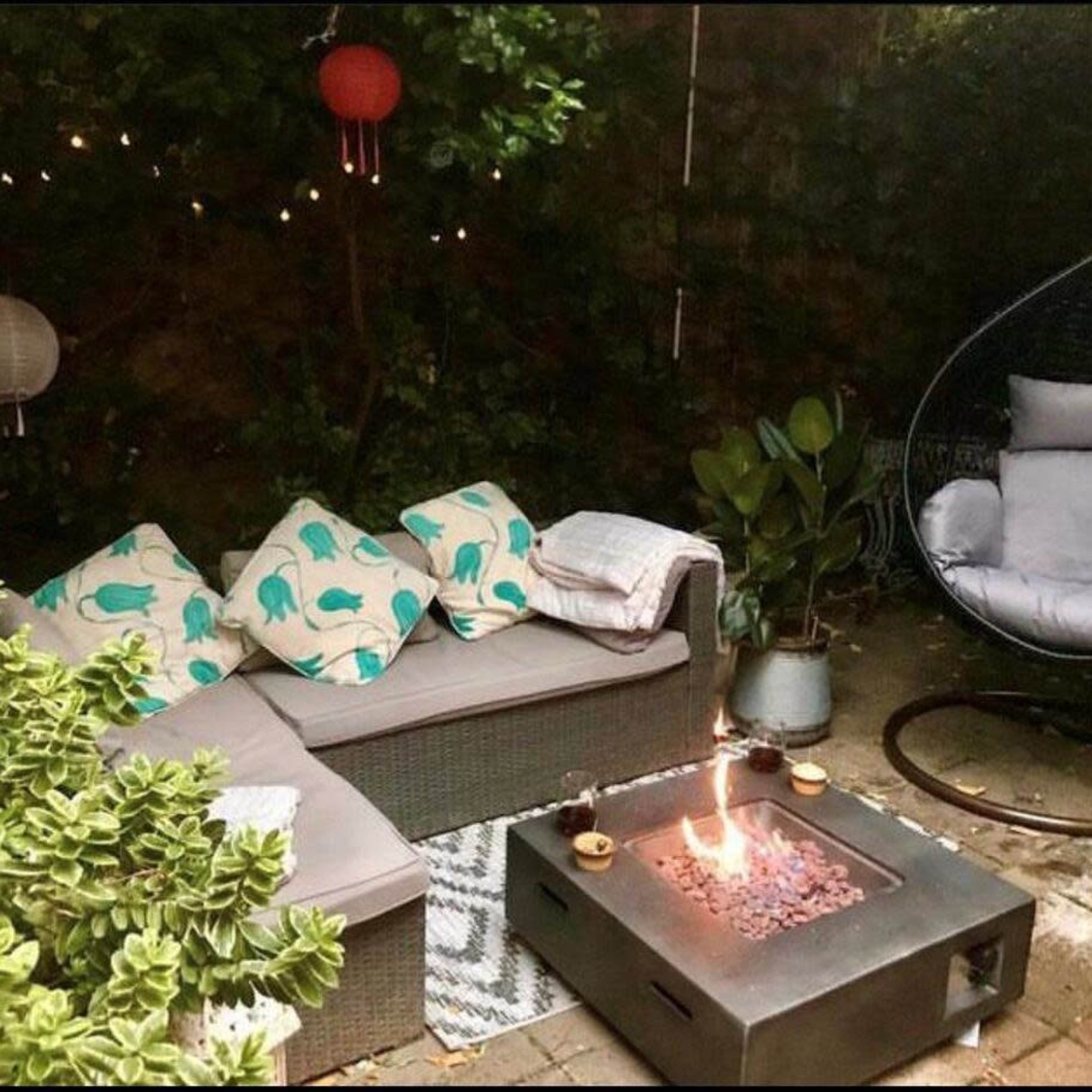 A cozy outdoor seating area with a sectional couch, a hanging chair, and a fire pit surrounded by greenery and decorative lanterns.