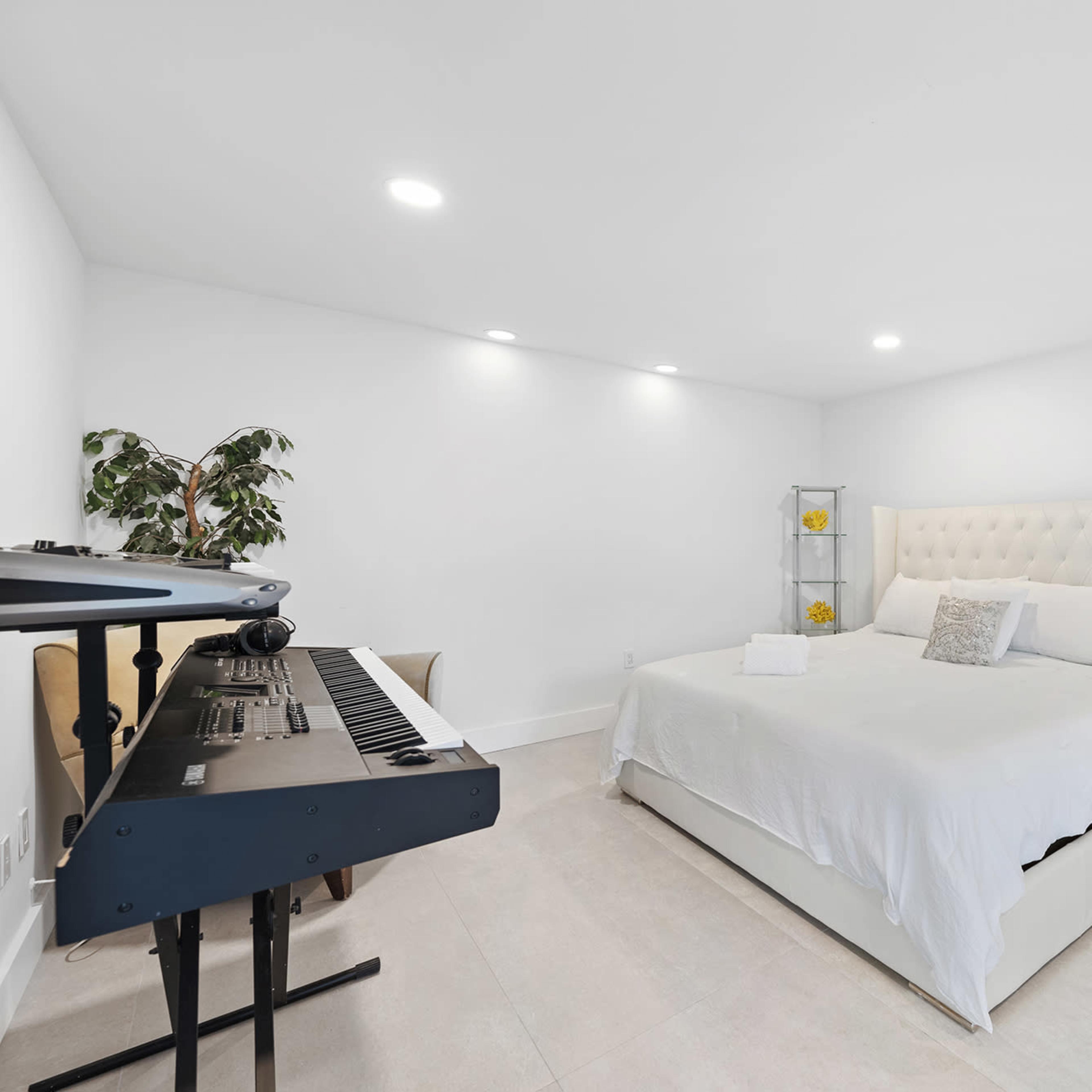 A minimalist bedroom featuring a large white bed, a keyboard, a plant, and a standing mirror.