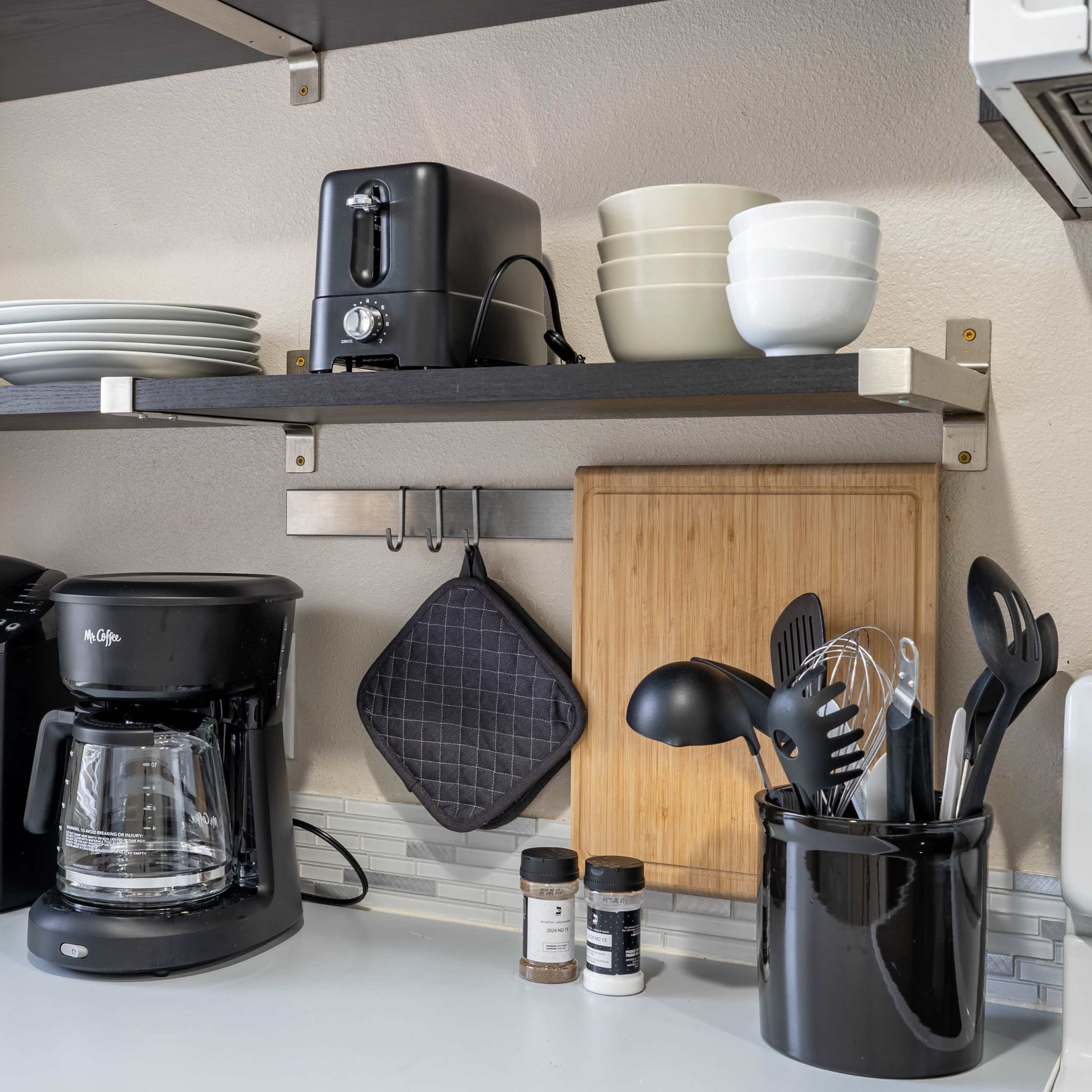 A kitchen countertop features a coffee maker, a toaster, and a coffee grinder, with neatly stacked plates and bowls on shelves above.